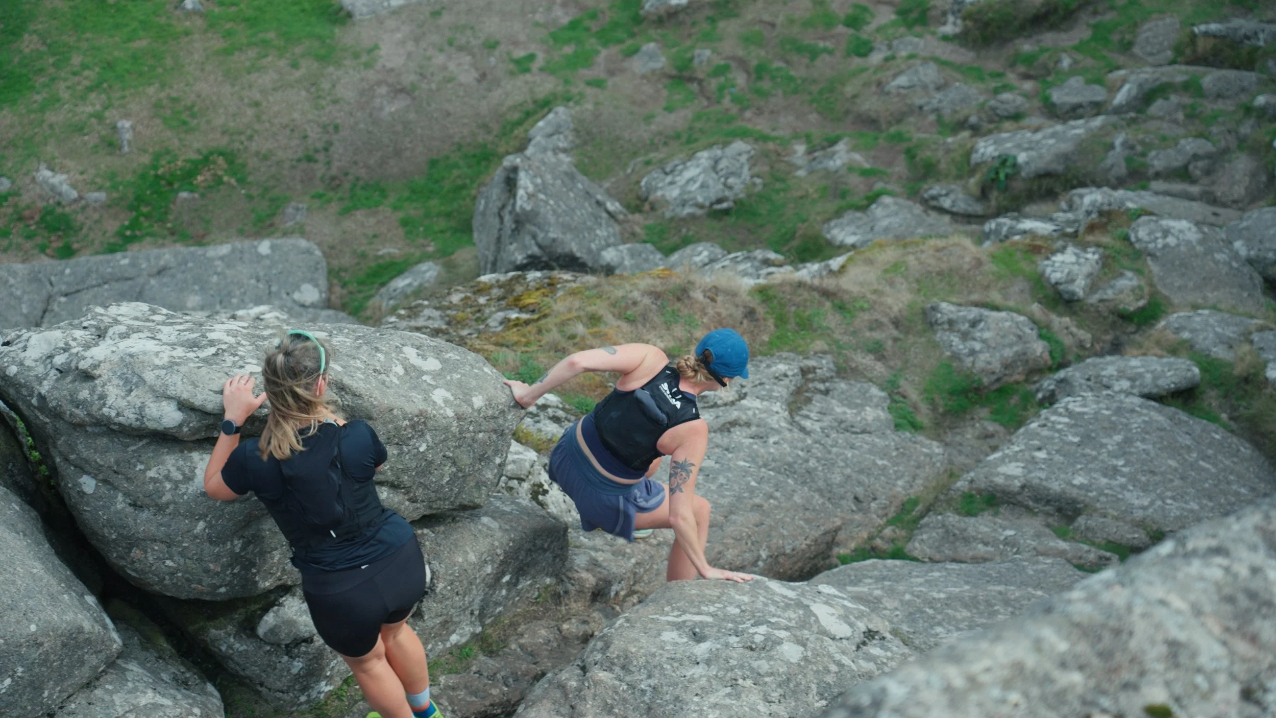 Two women rock climbing on a steep outdoor rock face with a grassy hillside in the background.