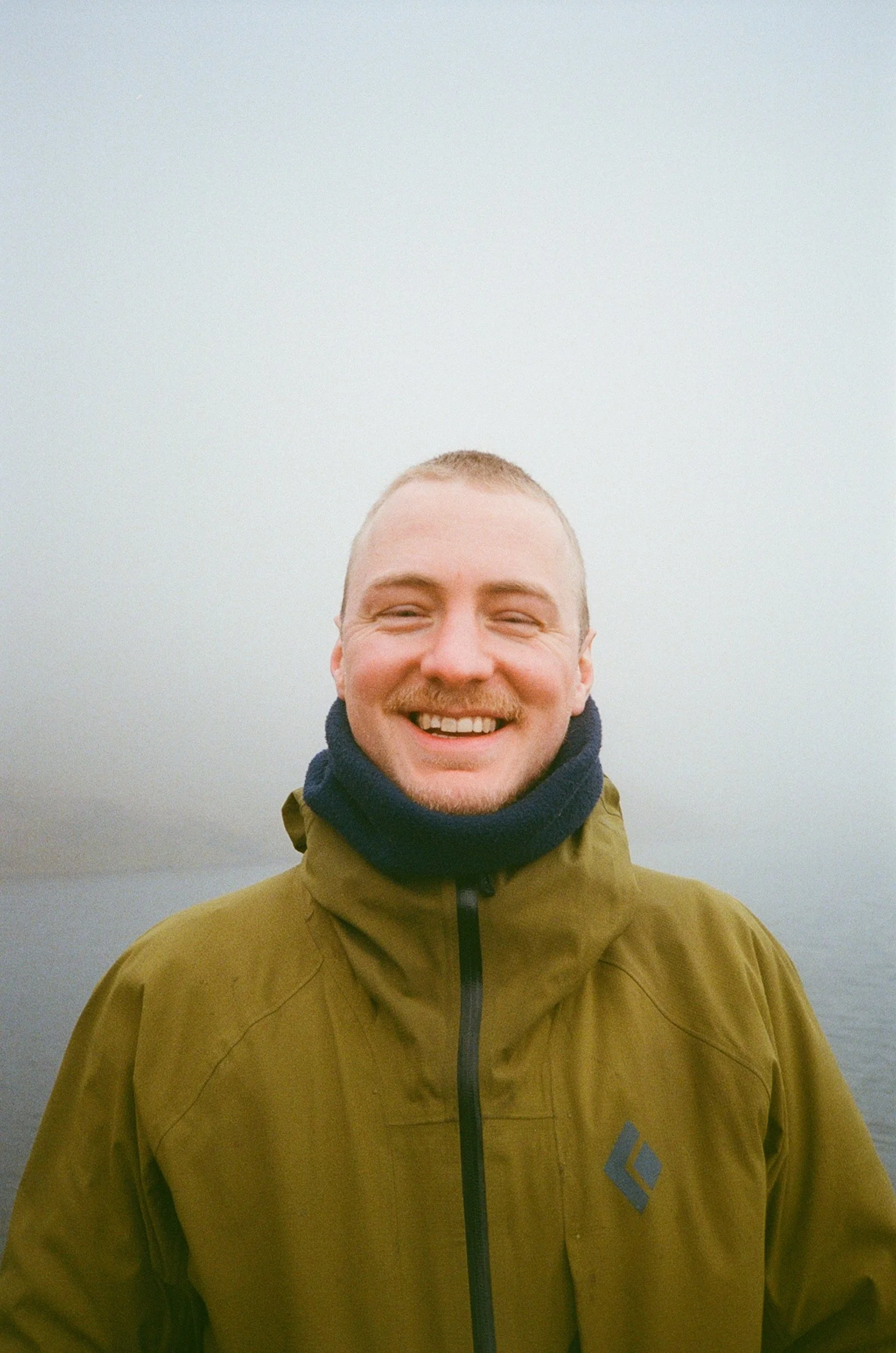 A smiling man with short hair and a beard, wearing a green outdoor jacket, standing against a foggy or misty background in Wales.