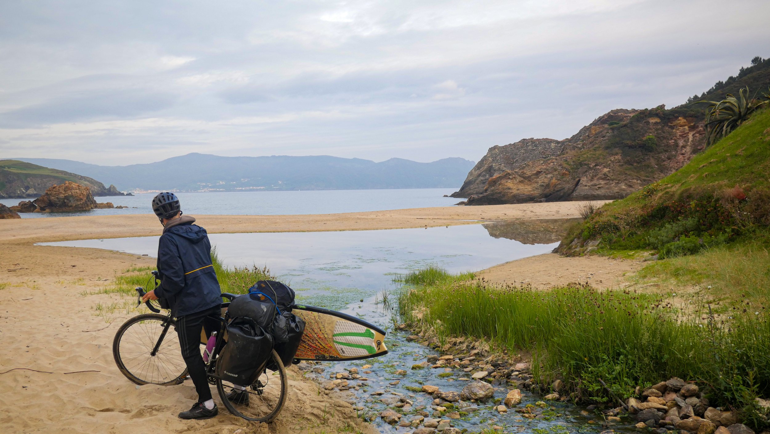 A person wearing a helmet and dark jacket standing with a bicycle and surfboard on a sandy beach in Northern Spain with rolling water, rocks, and green vegetation, overlooking a body of water with mountains in the background under a cloudy sky.