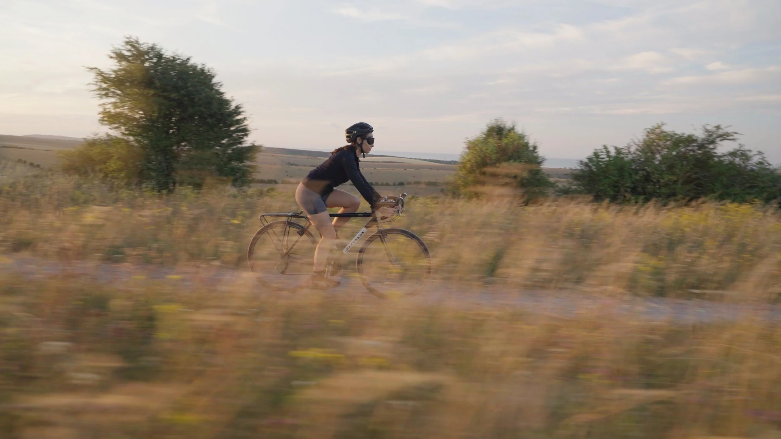 A woman wearing a helmet and sunglasses riding a bicycle through a rural landscape with trees and open fields during daylight.