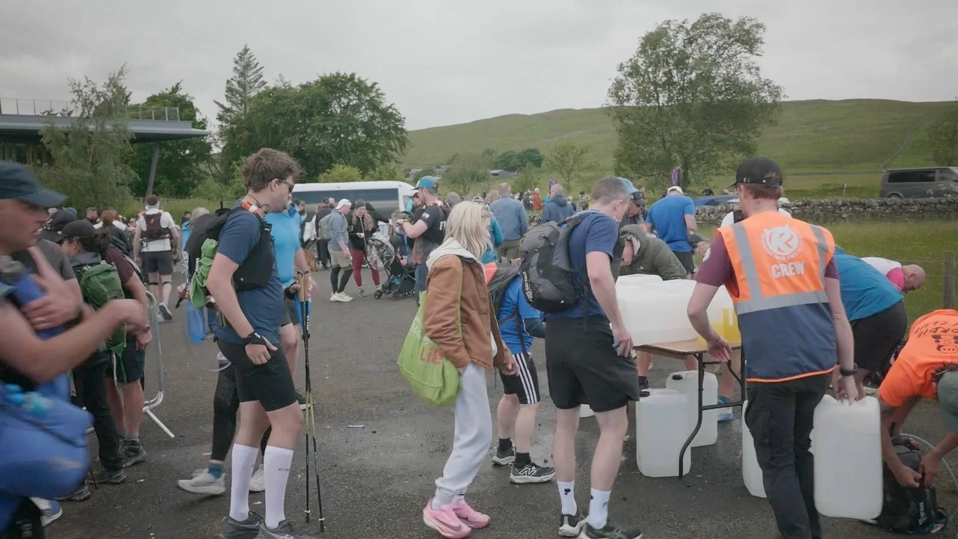 Crowd of runners at a check point in a rural area with rolling hills and trees in the background. Many people are with backpacks, some are interacting at tables, and a staff member in an orange vest marked 'Crew' is assisting.