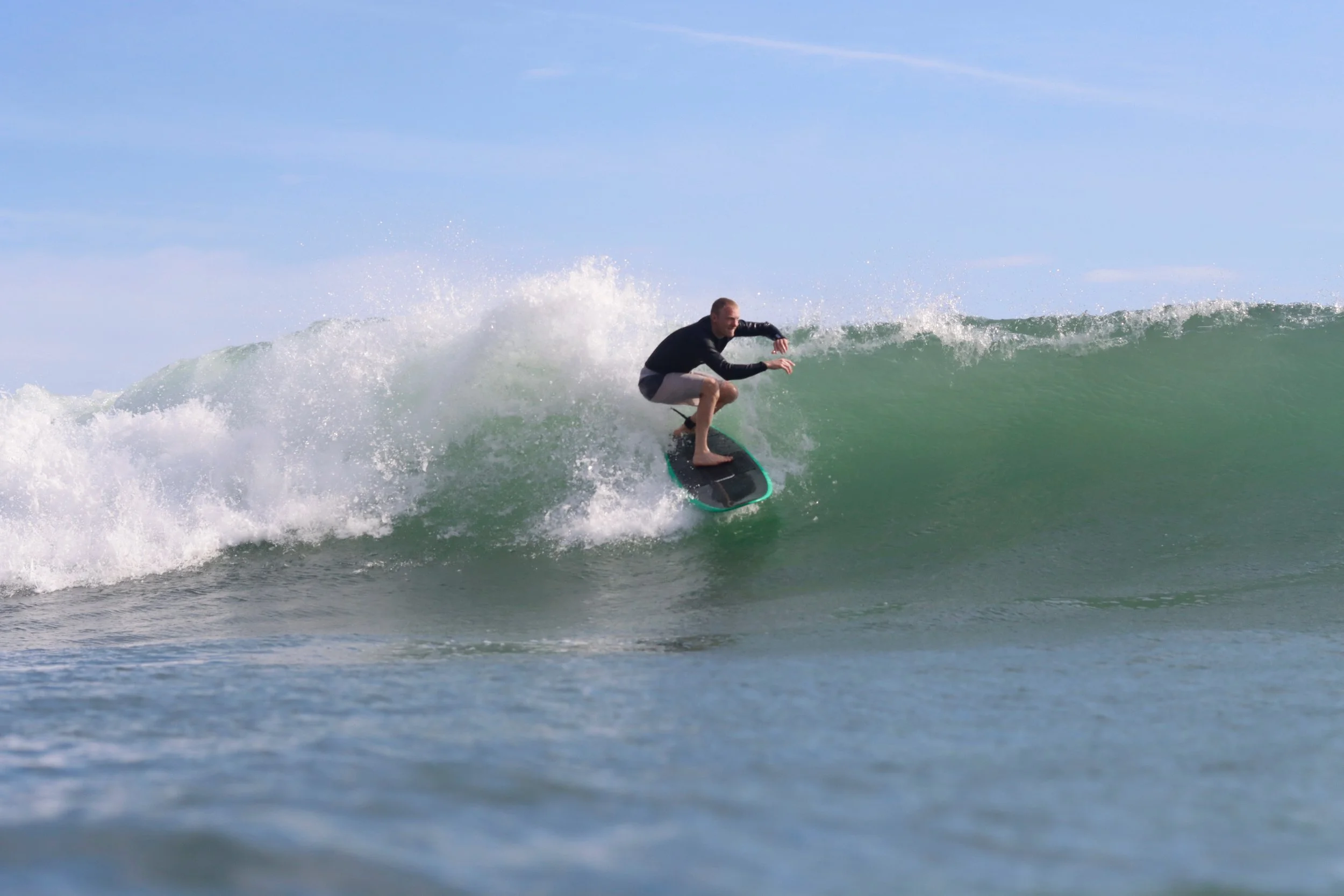 A man surfing on a wave in the ocean, wearing a black wetsuit and gray shorts, with a blue sky in the background.