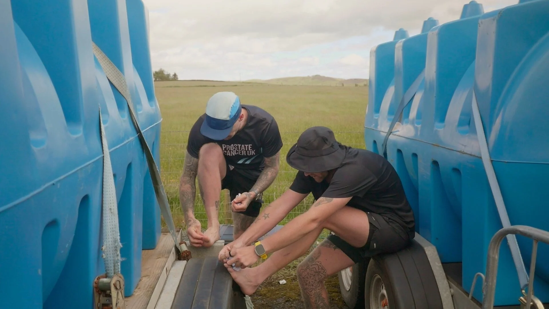 Two young men are putting plasters on blisters during an ultramarathon in a grassy field, with cloudy skies above.