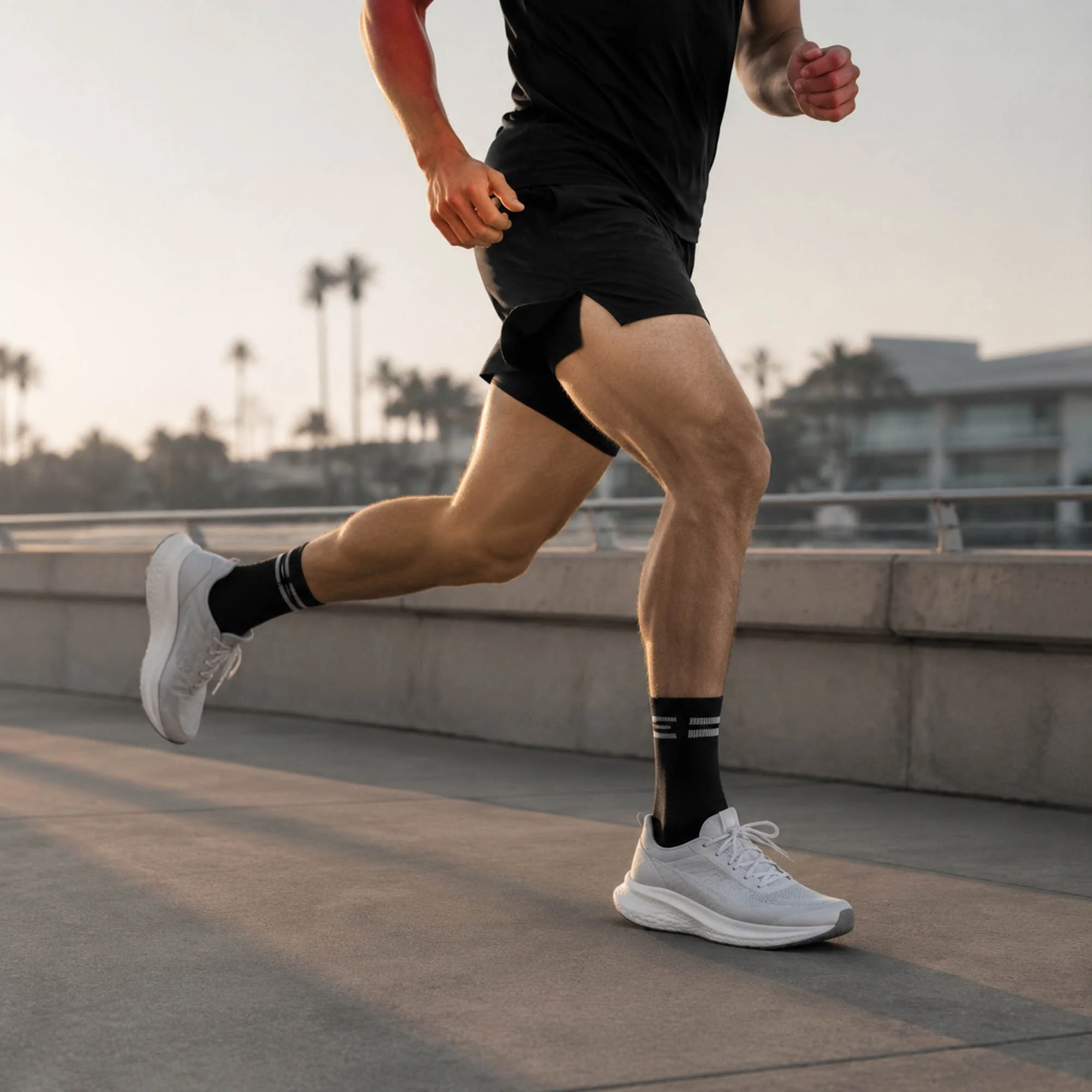 A man running outdoors on a concrete surface wearing black athletic shorts, a black shirt, white running shoes, and black socks with white stripes, with palm trees and buildings in the background during sunset.