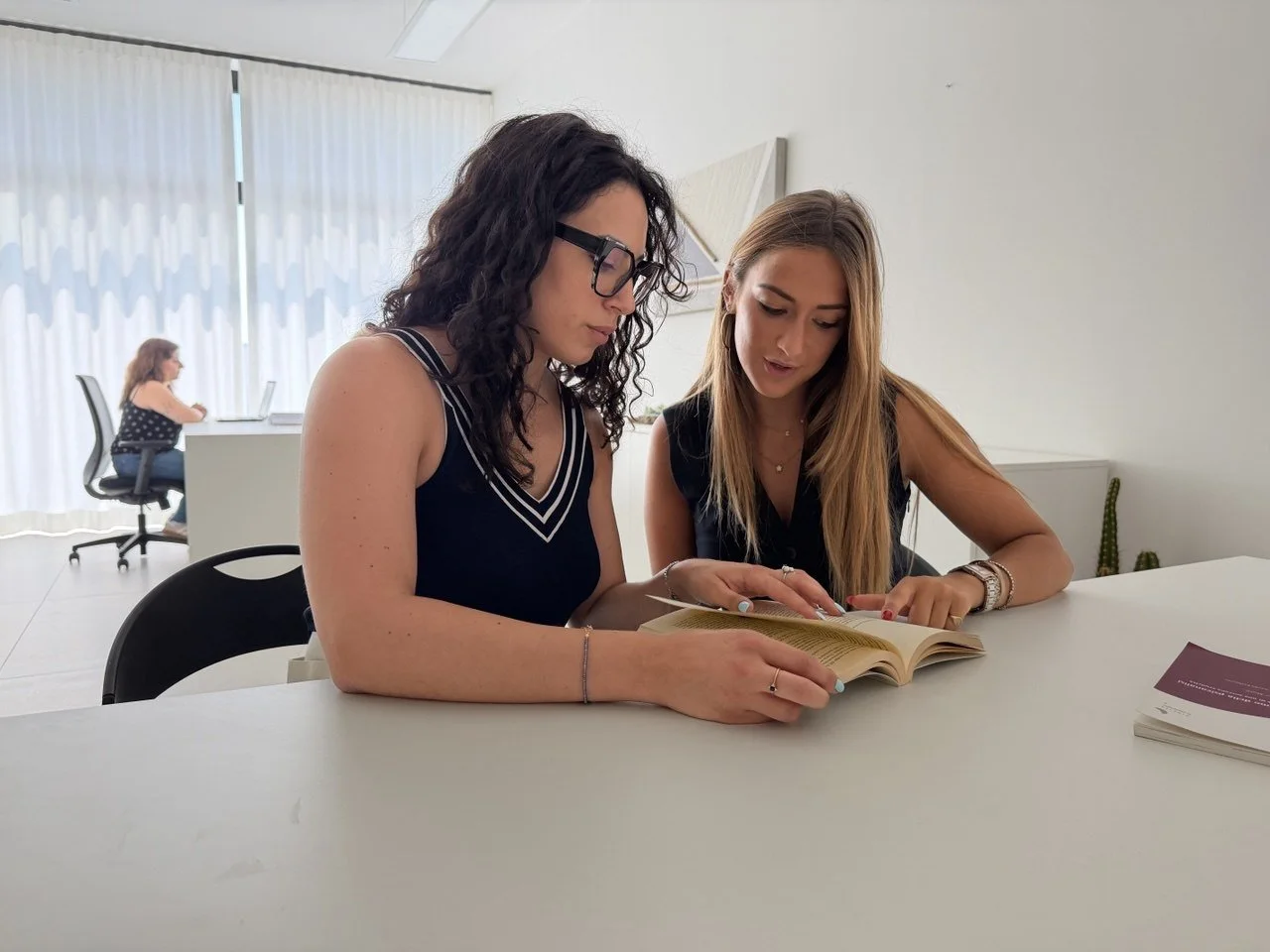 Due donne sedute a un tavolo che leggono un libro insieme in un ufficio moderno, con una donna che lavora al computer sullo sfondo.