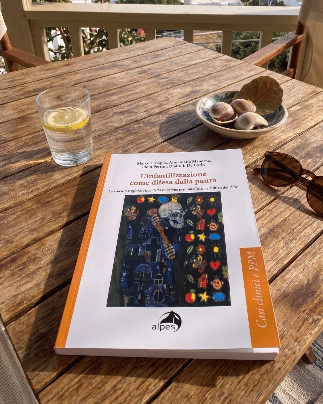 Tavola di legno con un libro intitolato "L'infantilizzazione come difesa dalla paura", un bicchiere di acqua con limone, un piatto con conchiglie e una foglia, un paio di occhiali da sole e una finestra sullo sfondo.