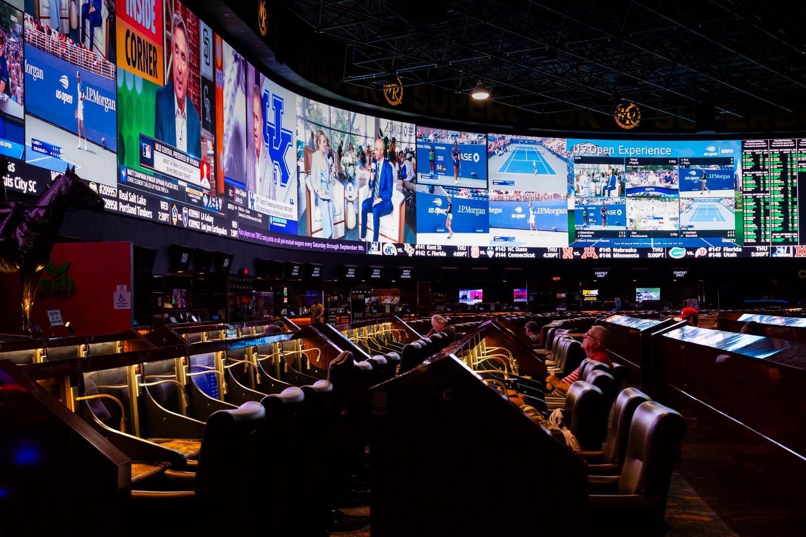 A sports betting lounge at an airport with large display screens showing tennis matches, news, and advertisements, and people sitting in leather chairs watching the screens.