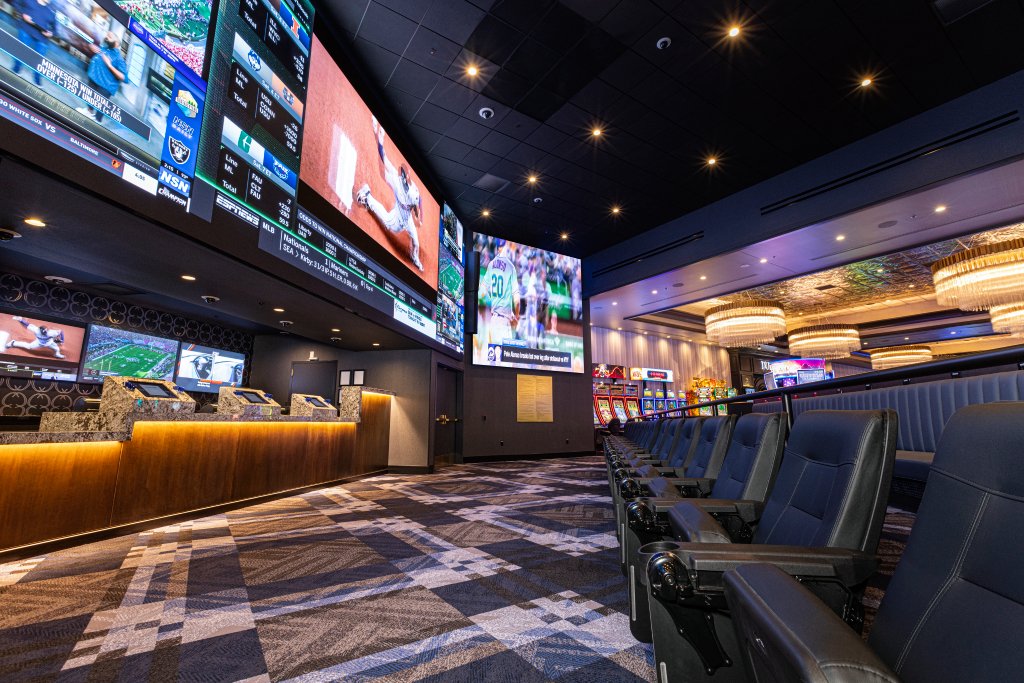 A dimly lit sports bar with multiple large screens displaying sports games, a row of empty black chairs facing the screens, and a bar counter with electronic kiosks, with decorative chandeliers hanging from the ceiling.