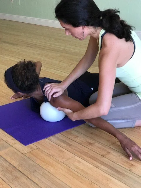 A woman assisting a young girl with a yoga pose on a purple mat, using a white yoga block for support in a room with wooden flooring.