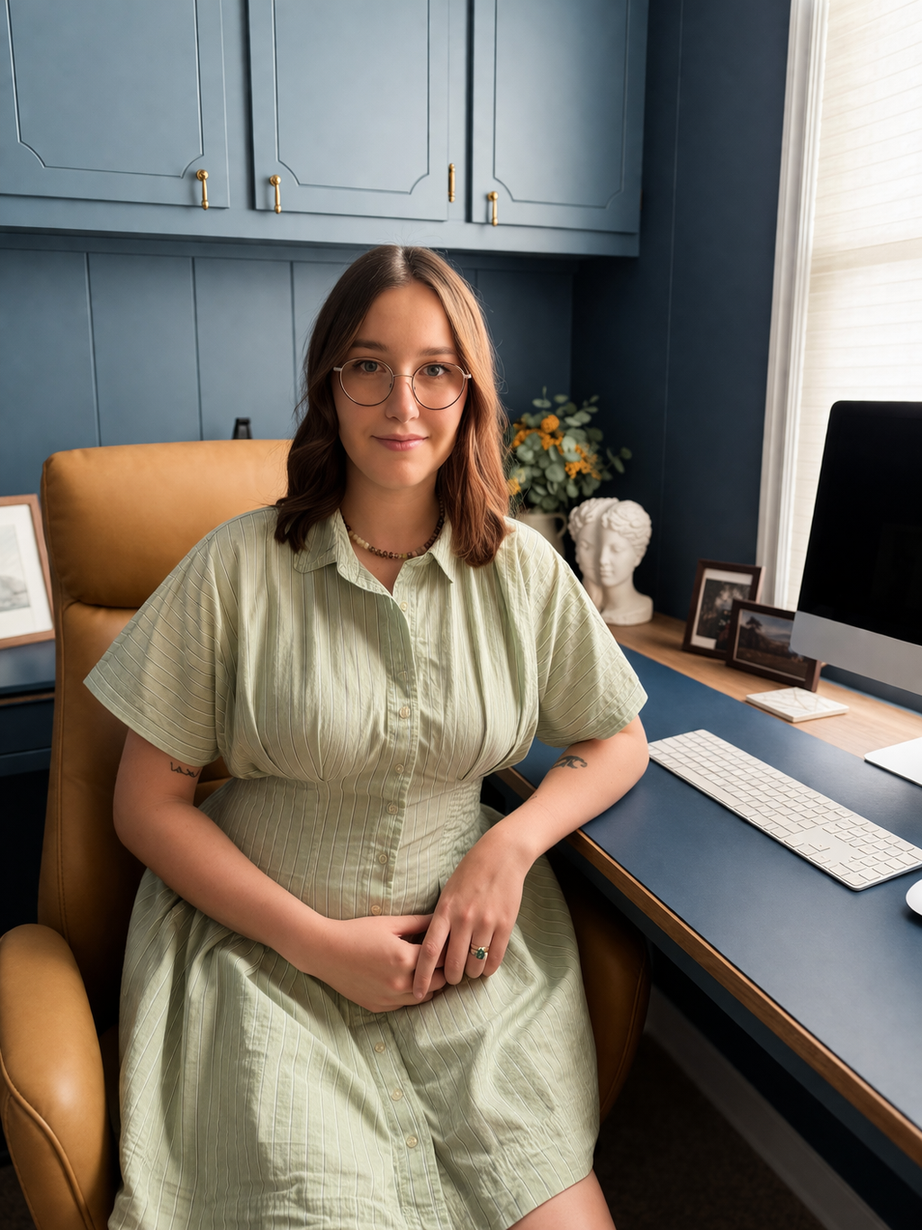 A young woman with glasses and shoulder-length brown hair, wearing a pale green striped dress, sits in a mustard-yellow chair in a home office, with a computer, framed photos, flowers, and a white bust in the background.