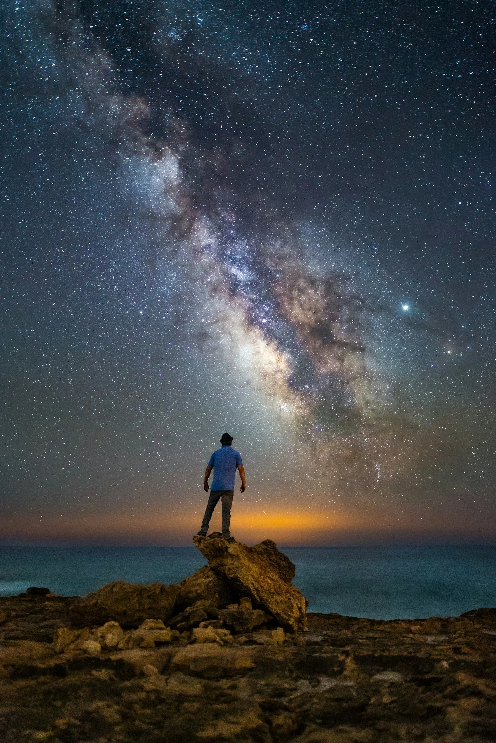 A person standing on rocks next to the ocean, looking at the night sky filled with stars and the Milky Way galaxy.