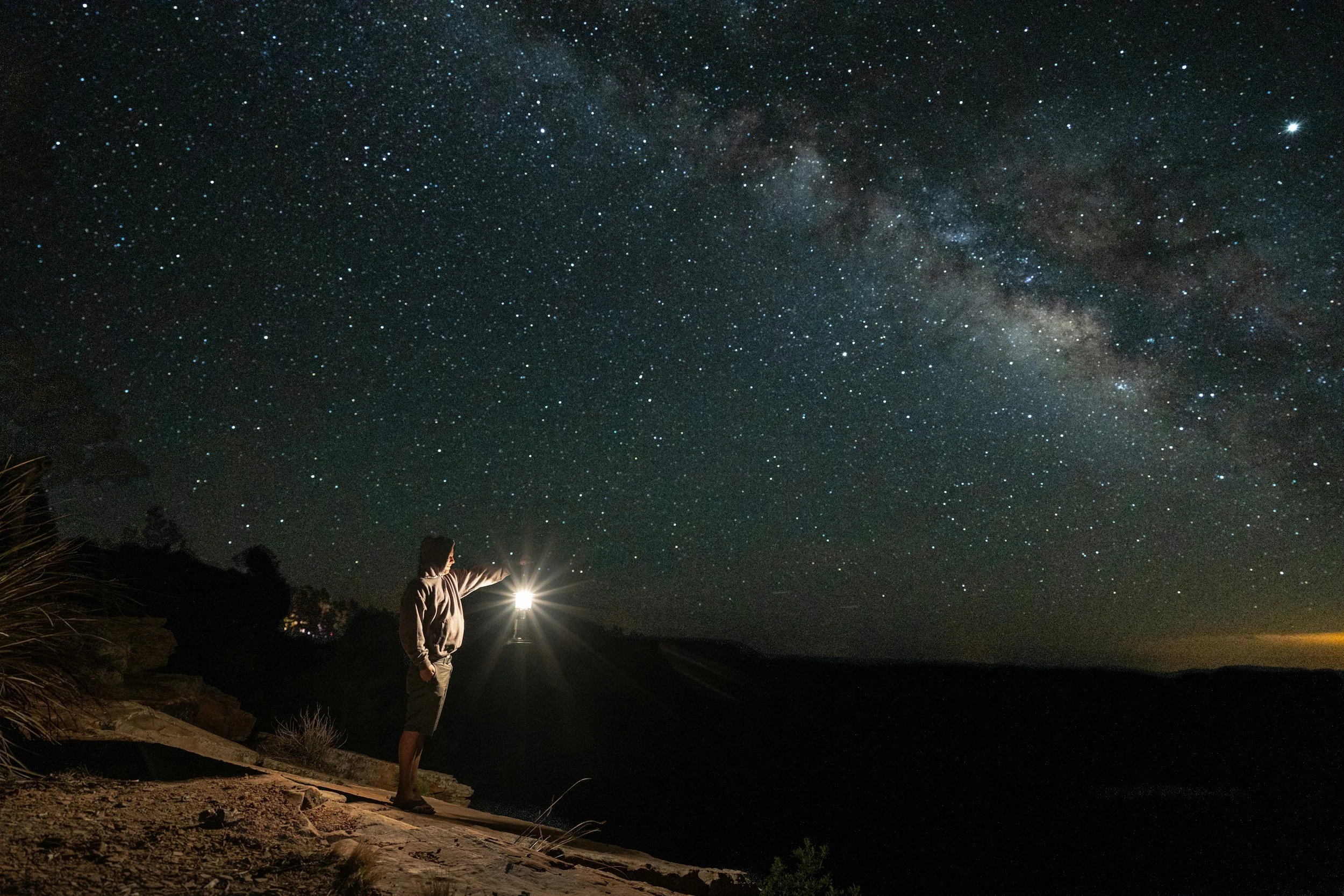 A person holding a flashlight pointing towards the starry night sky and the Milky Way galaxy, standing on a rocky shoreline, as if seeking the answer to a question.