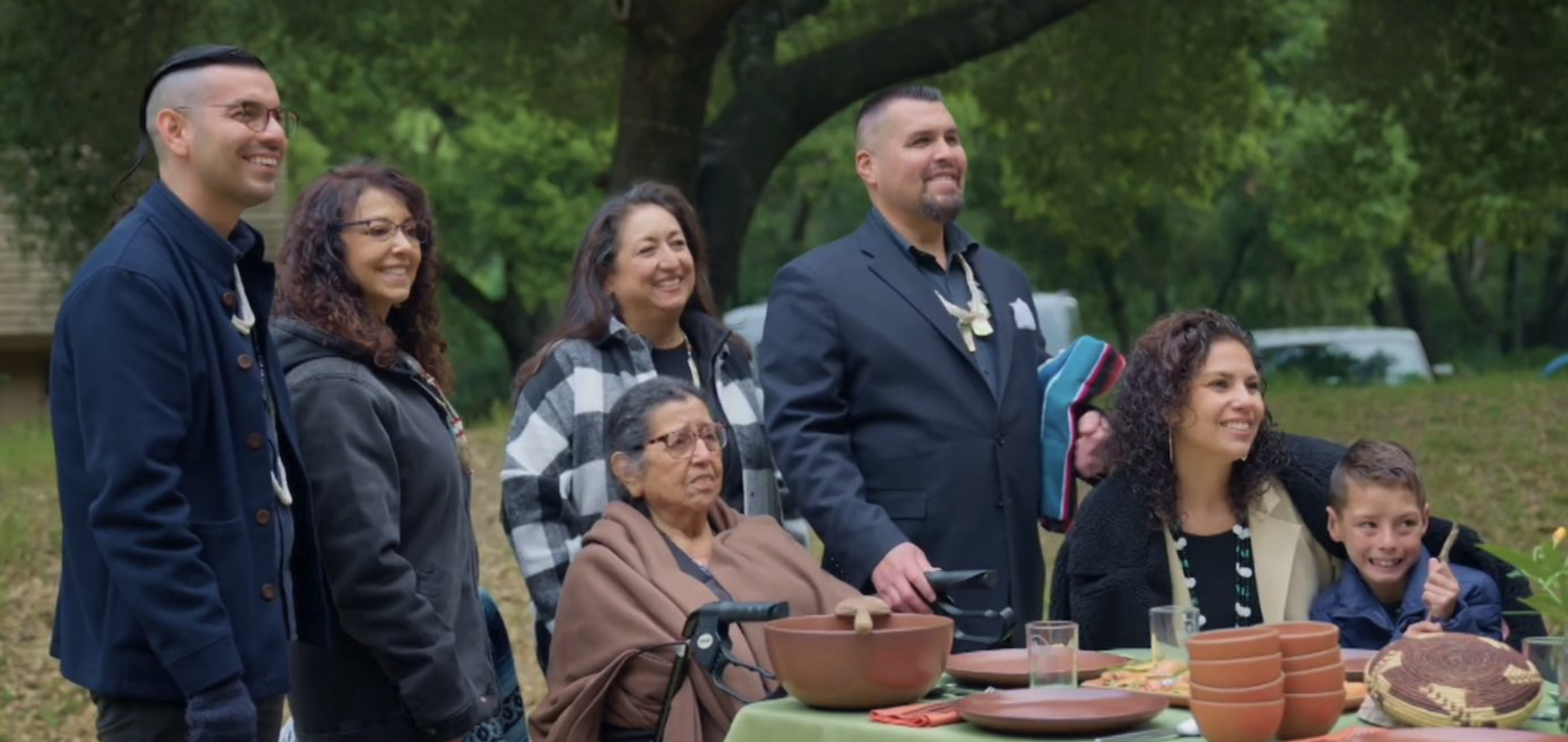 Group of diverse people smiling outdoors at a picnic table with pottery and bowls.