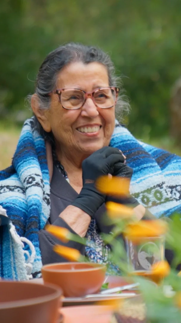 Smiling elderly woman with glasses and gray hair, wearing a dark cardigan and a colorful shawl, sitting outdoors at a table with ceramics, surrounded by greenery.
