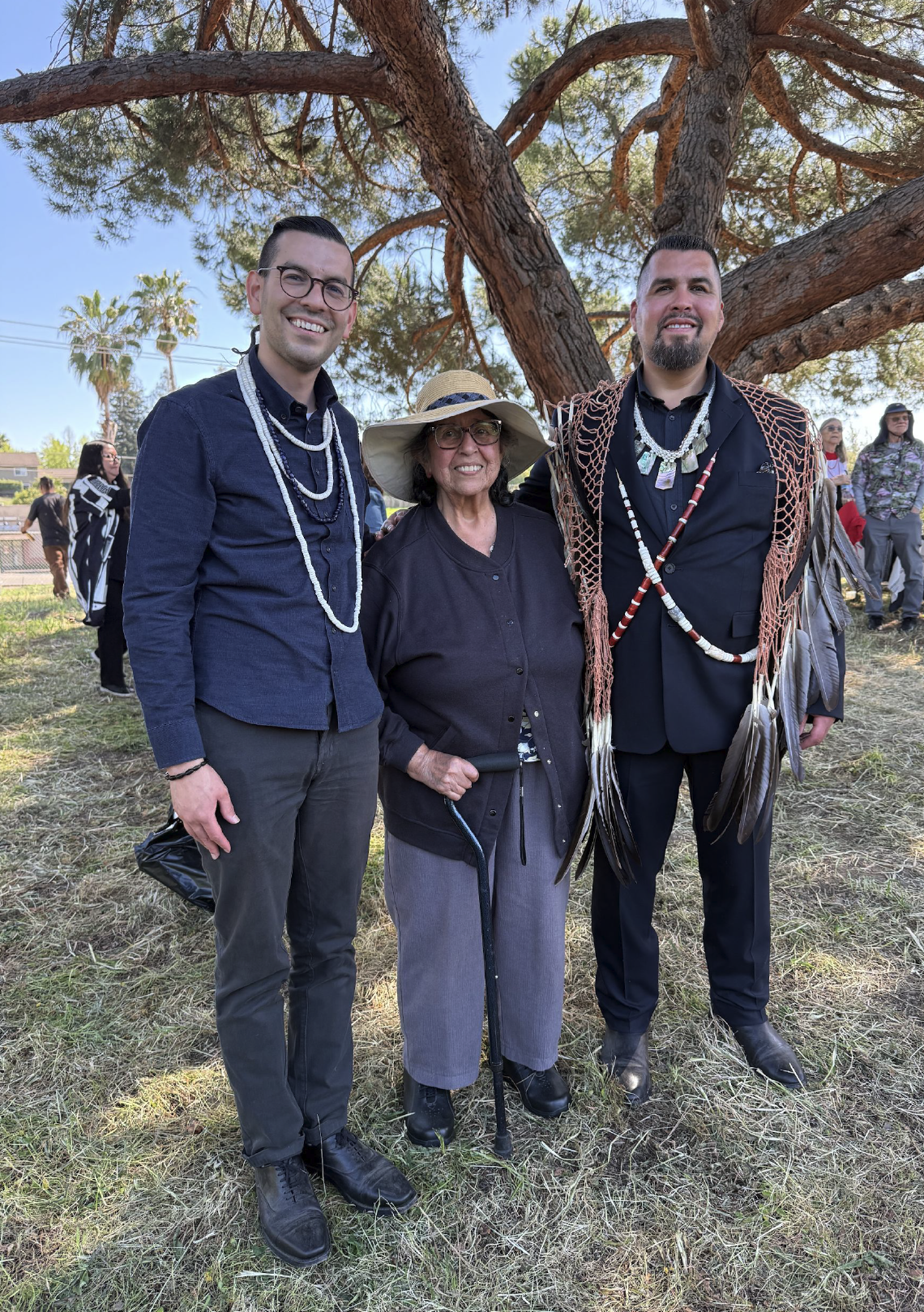 Three people standing outdoors under a large tree, smiling at the camera. Two men are wearing dark suits with Native American jewelry, and a woman in the middle wears a wide-brimmed hat, glasses, and a dark cardigan. Other people can be seen in the background.