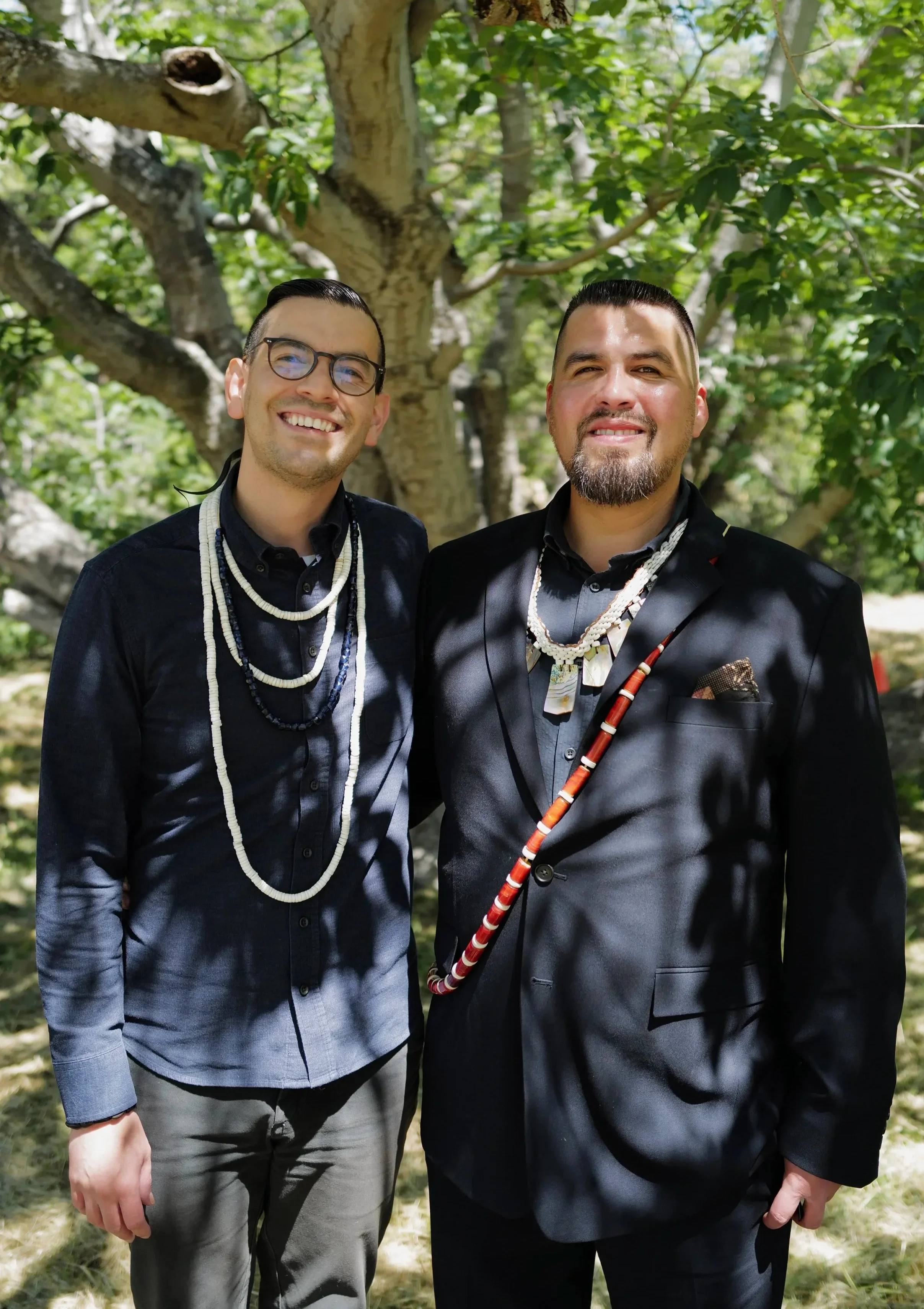 Two men standing outdoors in front of a tree, smiling at the camera. They are dressed in formal attire with necklaces, and one is holding a walking stick.