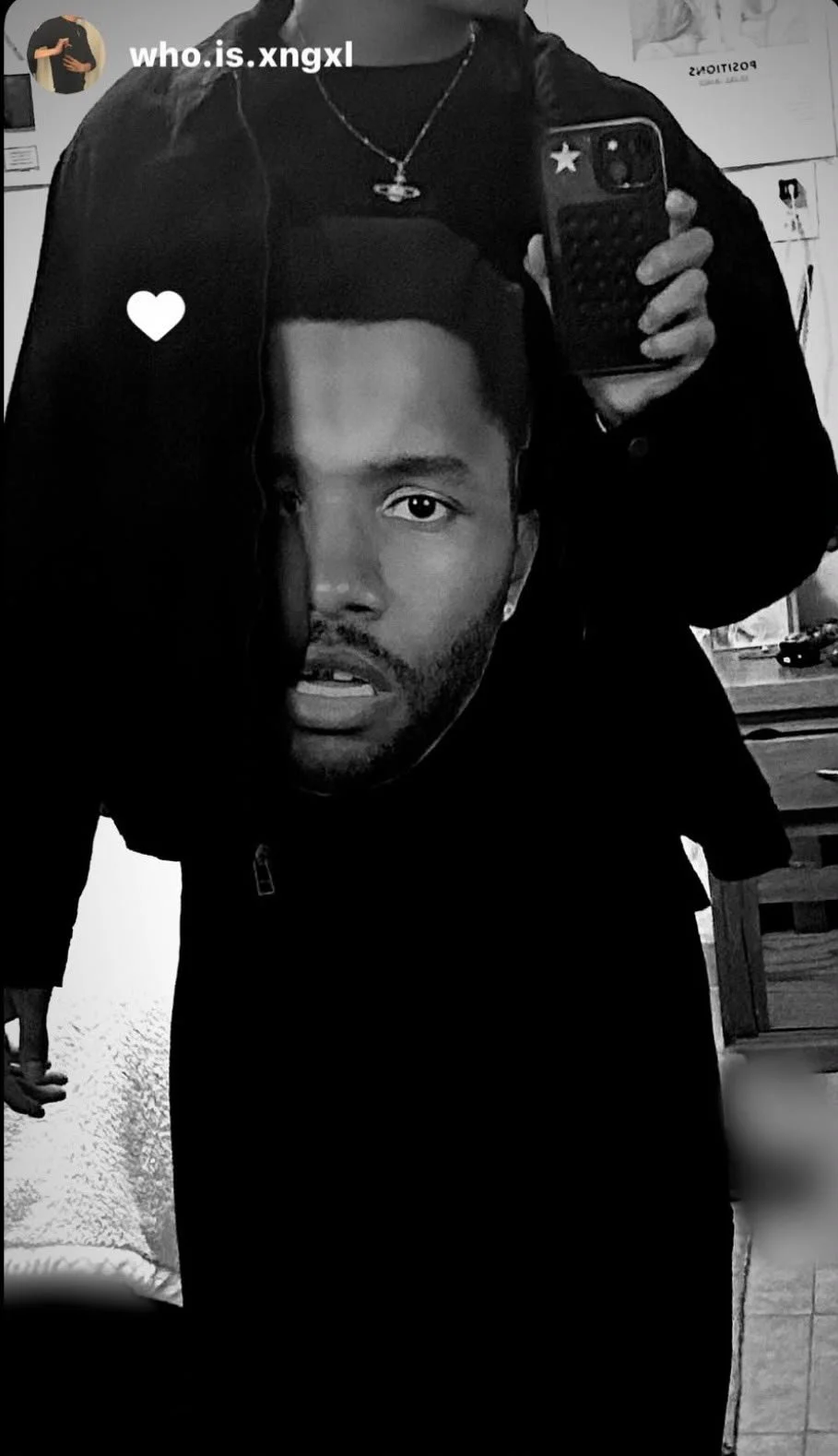 A person taking a mirror selfie in black and white, wearing a hoodie with a large printed face on it, a necklace, and holding a phone with a star-shaped sticker on the case.