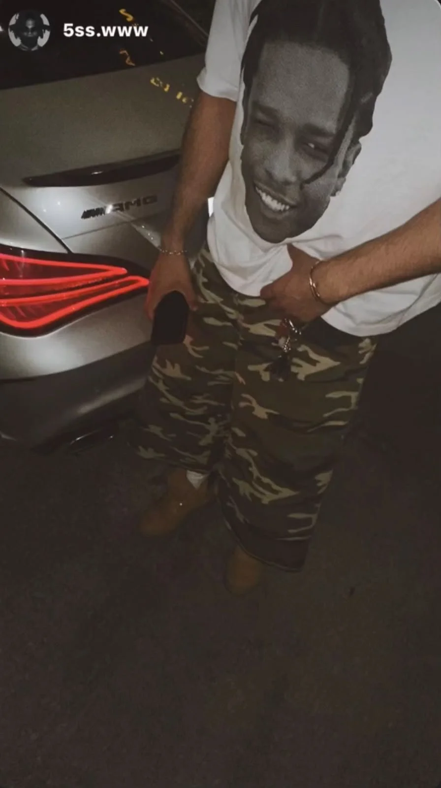 A man standing next to a silver Mercedes-Benz car at night, smiling and dressed in a white t-shirt with a graphic print, camouflage shorts, and brown shoes. He has a gold bracelet and a ring on his right hand.