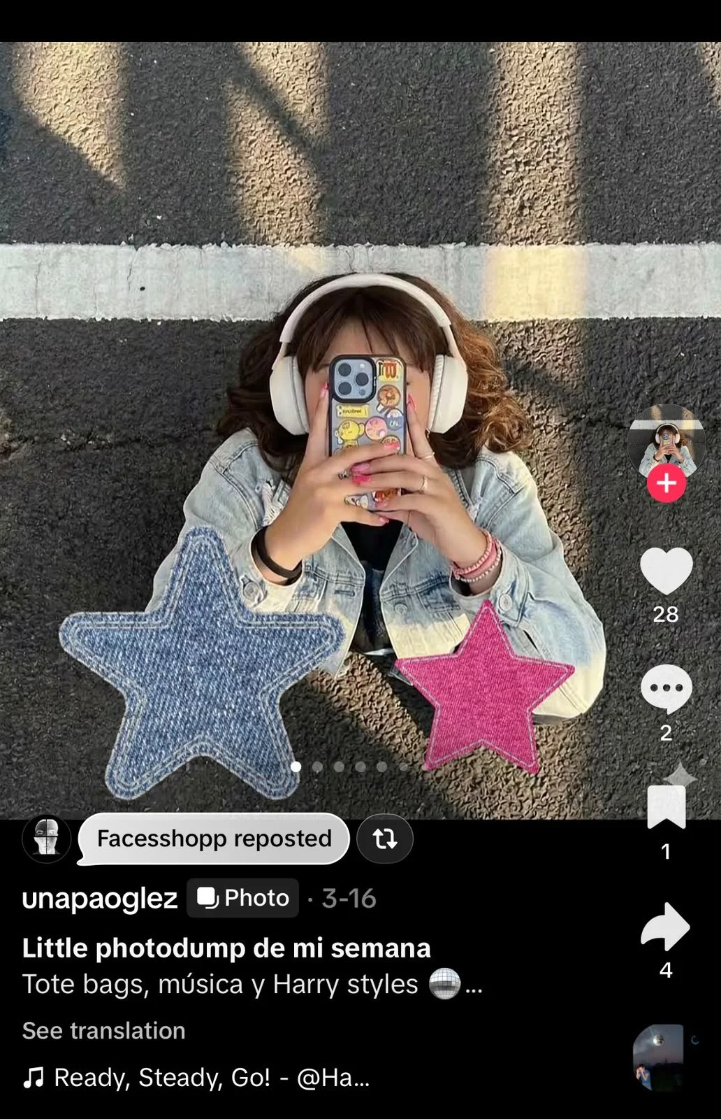 A young woman lying on the ground, taking a selfie with her smartphone. She is wearing large headphones and a denim jacket, with two large star-shaped plush toys, one blue and one pink, on either side of her head. The ground appears to be asphalt with a white stripe running across the top of the image.