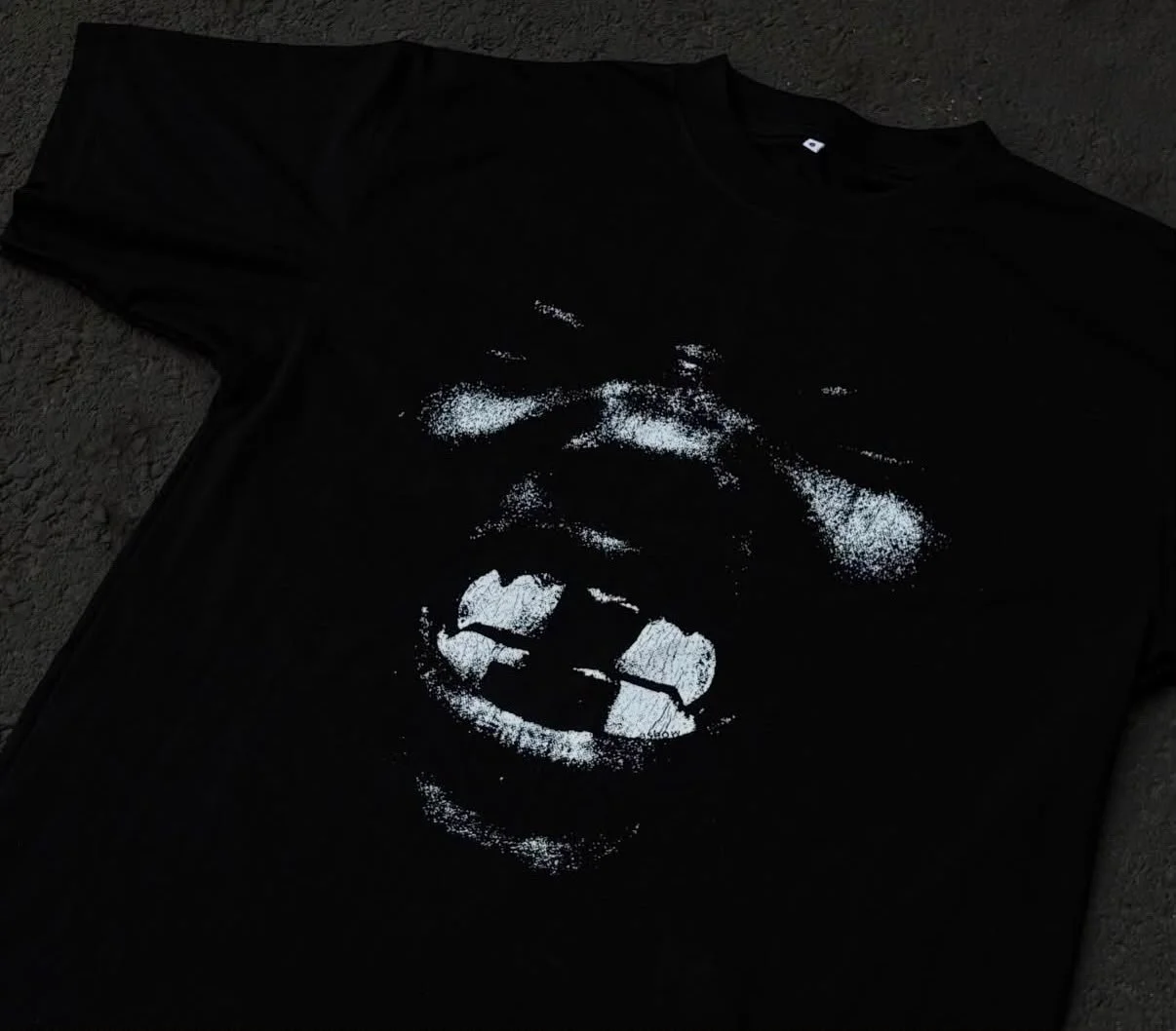 Black T-shirt with a white graphic of a person's face and open mouth, lying on a dark textured surface.