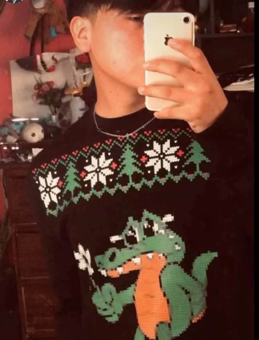 A person taking a mirror selfie wearing a black holiday sweater with white snowflakes, green Christmas trees, and a cartoon dinosaur holding a marshmallow roasting stick.