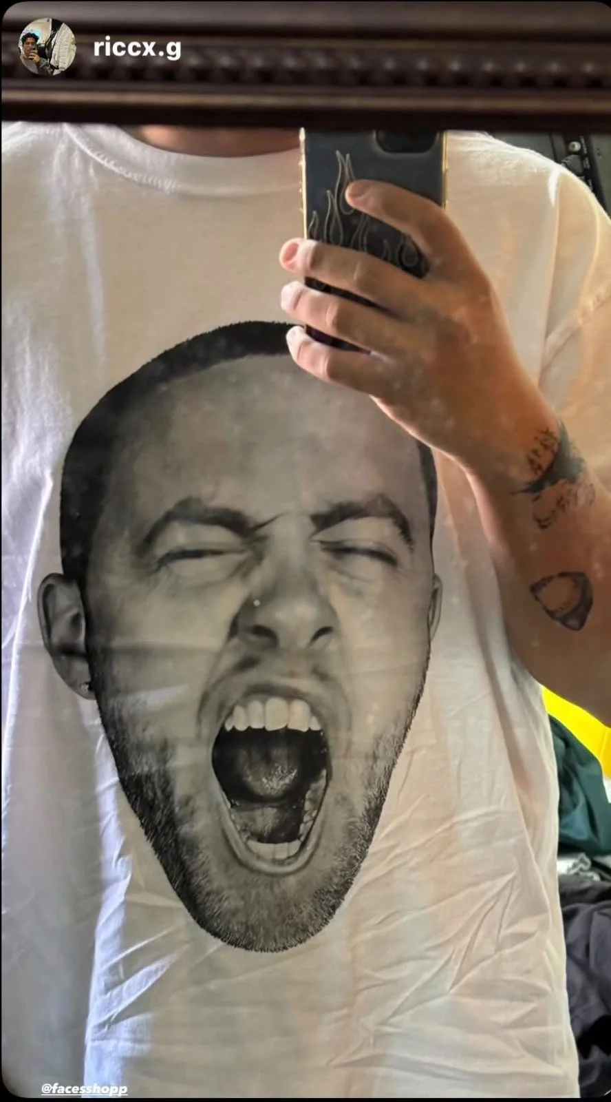 A person wearing a white T-shirt with a black-and-white graphic of a face screaming with eyes closed, mouth open, and teeth showing. The person's arm and hand are visible, holding a phone to take the photo in a mirror.
