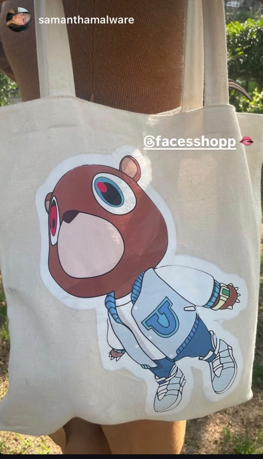 A tote bag with a cartoon bear character sticker, wearing a white jacket with a blue 'J' letter, blue hoodie, and sneakers, outdoors with green foliage in the background.