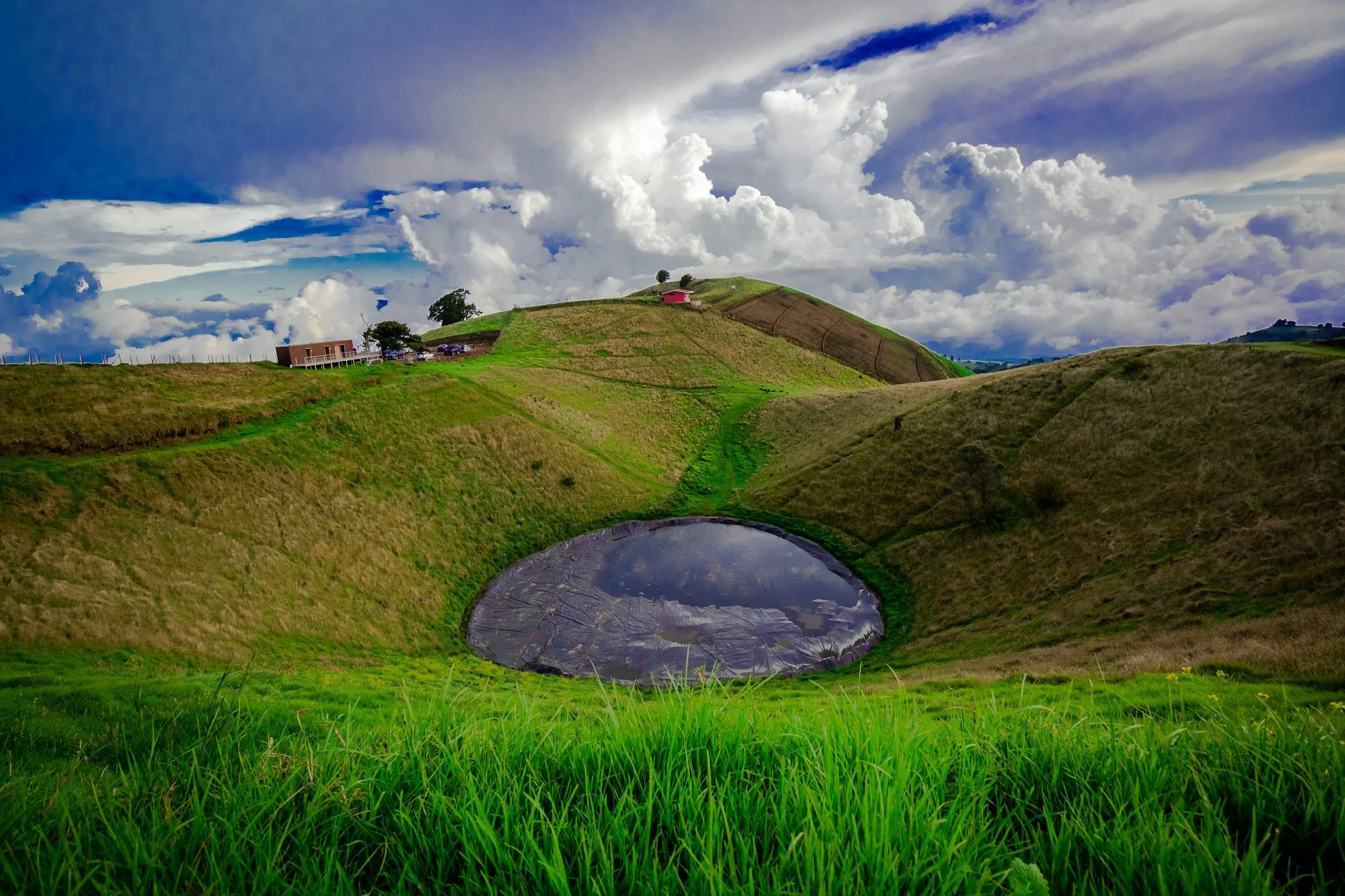 Green hilly landscape with a pond and farm buildings under a cloudy sky.