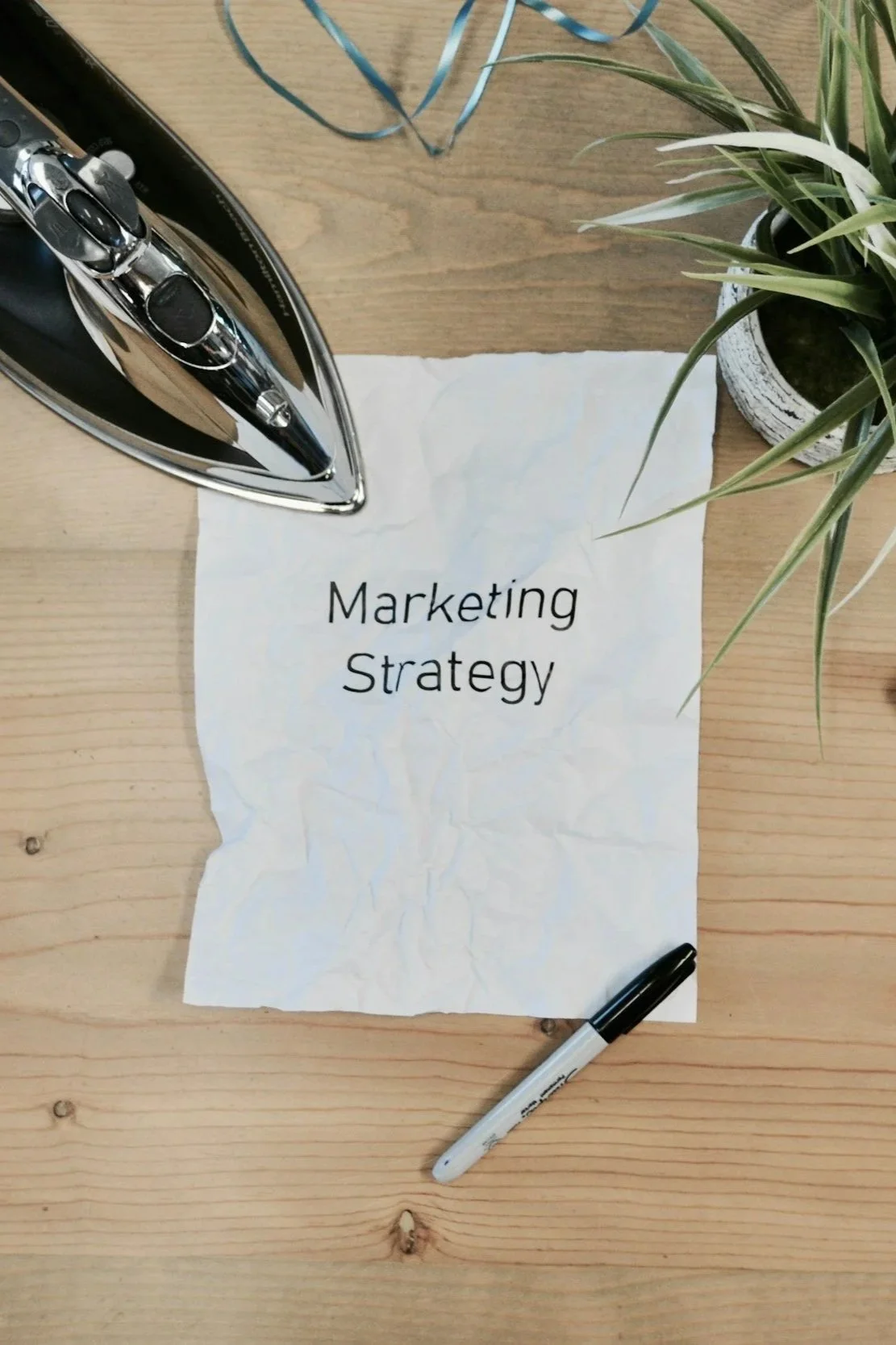 A crumpled piece of paper on a wooden desk with the words "Marketing Strategy" written on it. An ink pen is placed at the bottom of the paper. To the left, there is an iron and some blue ribbon on the desk. To the right, there is a potted plant with green leaves.