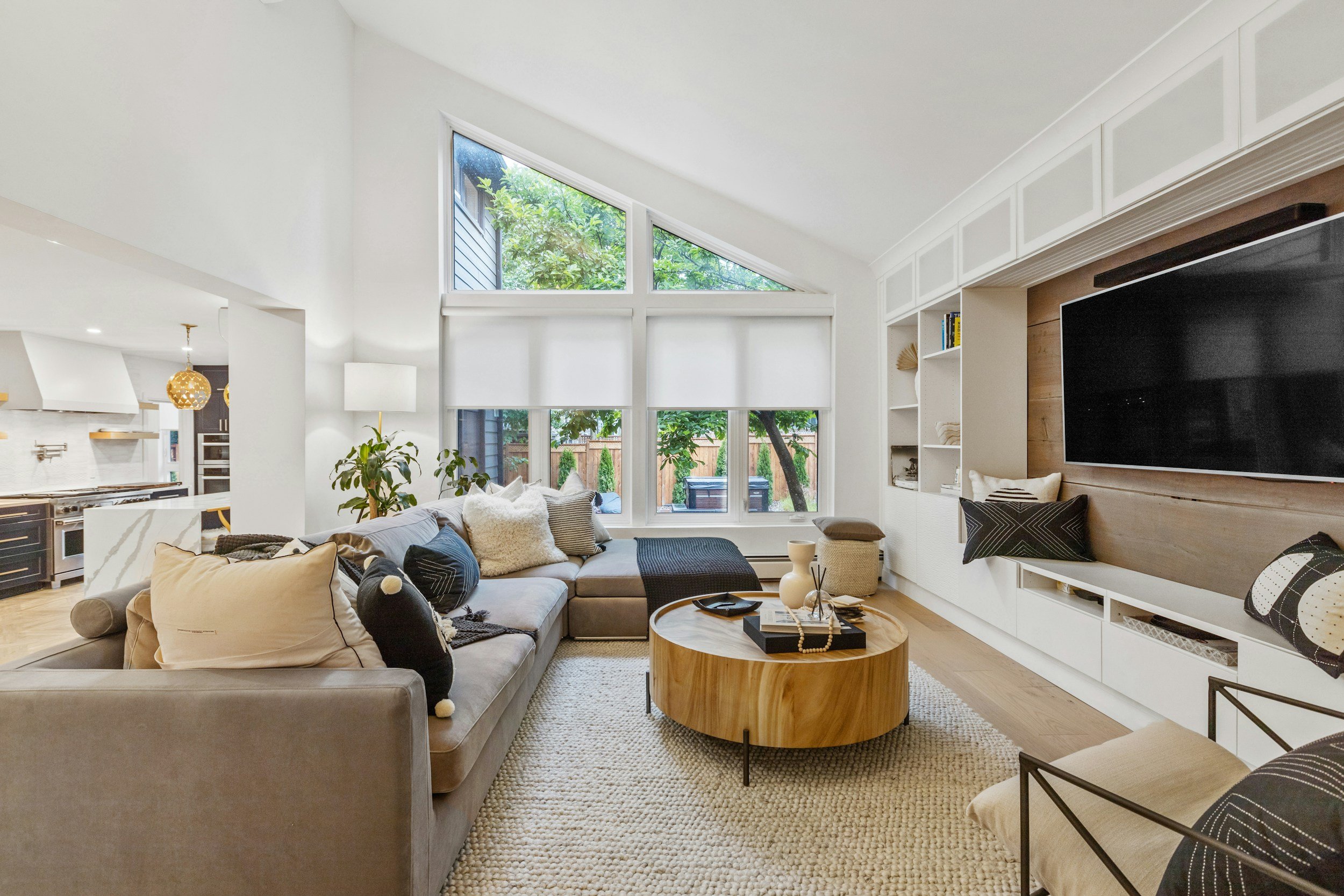 A modern living room with a large sectional sofa decorated with various pillows, a round wooden coffee table, a textured beige rug, a built-in white entertainment center with a flat-screen TV, and large windows revealing a green backyard.