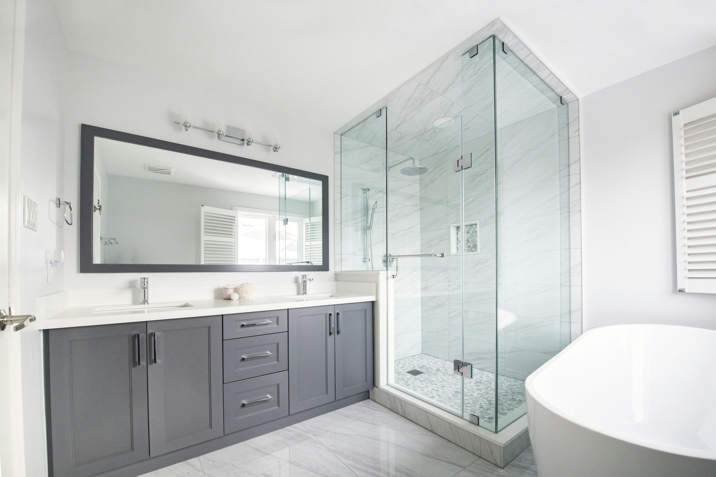 Modern bathroom with a glass-enclosed shower, a double vanity with dark gray cabinets, and a large mirror, with a white bathtub partially visible.