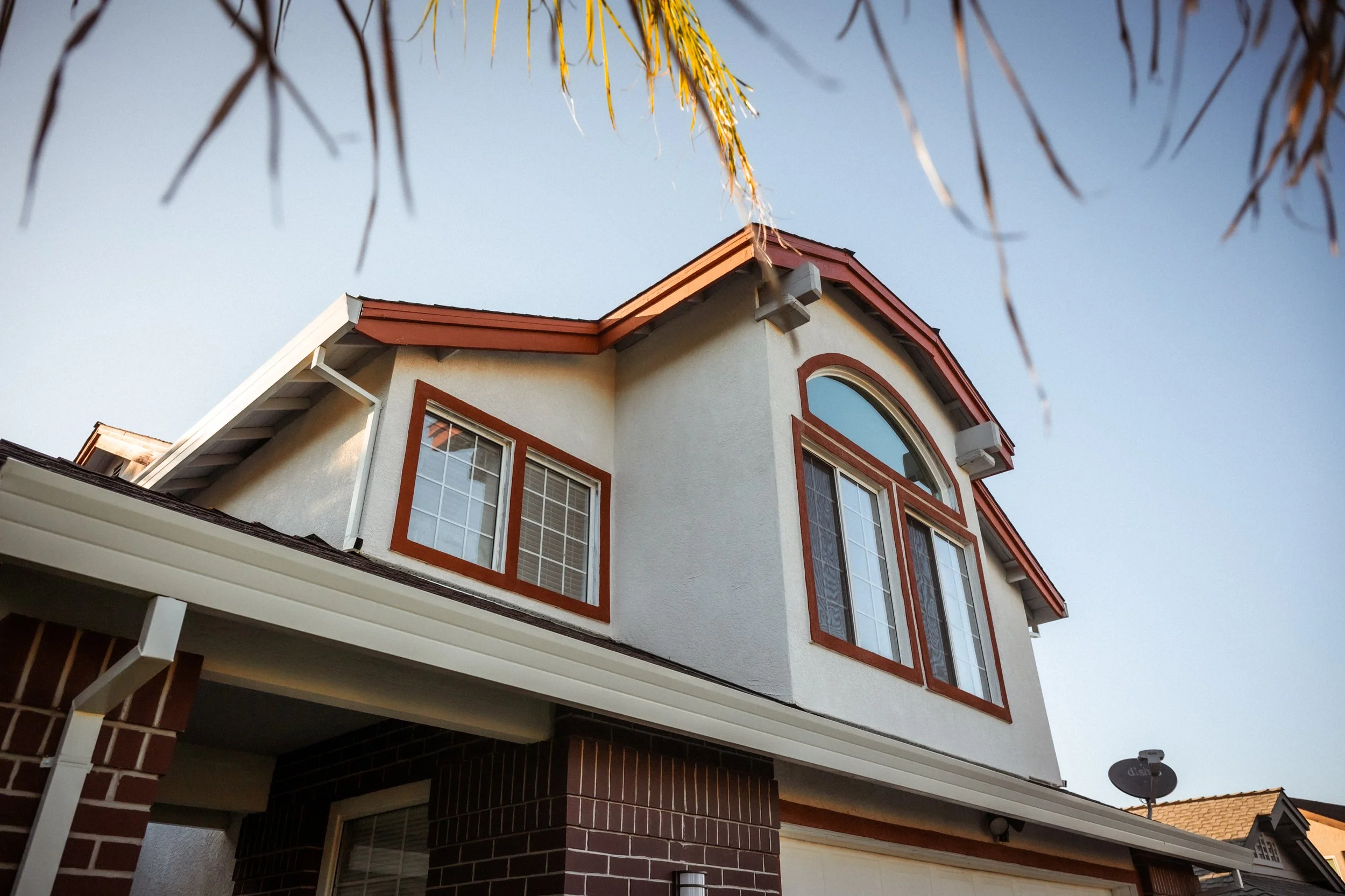 A multi-story house with a beige stucco exterior and red trim around the windows and roof. The house has large windows, an arched window at the top, and a satellite dish attached to the side. Tree branches with yellow leaves are visible above the hou