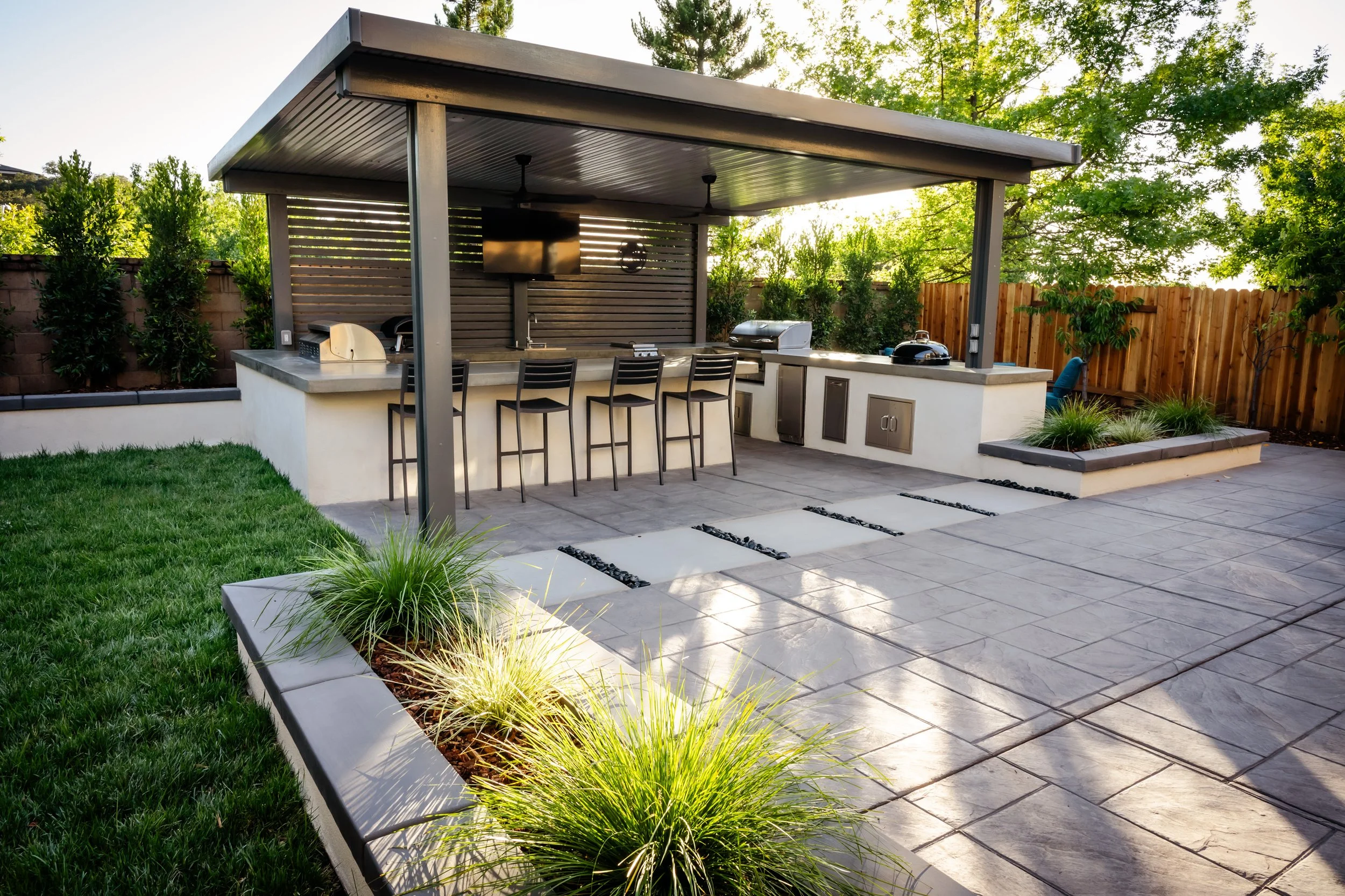 Backyard outdoor kitchen with a covered patio, barstools, grill, and plants, with sunshine and trees in the background.
