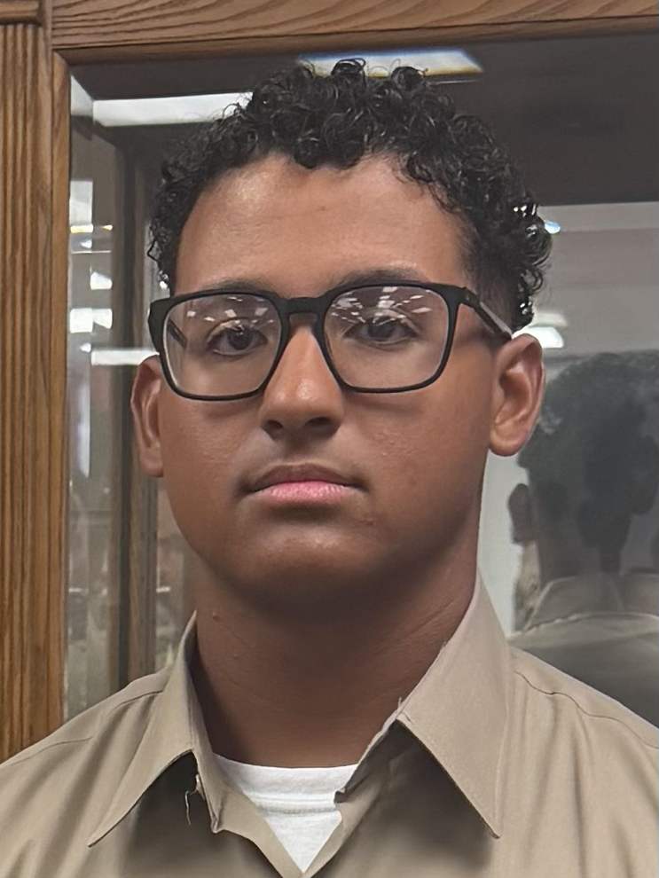 Young man with glasses wearing a beige uniform and white undershirt, standing in a room with wooden and glass display cases.