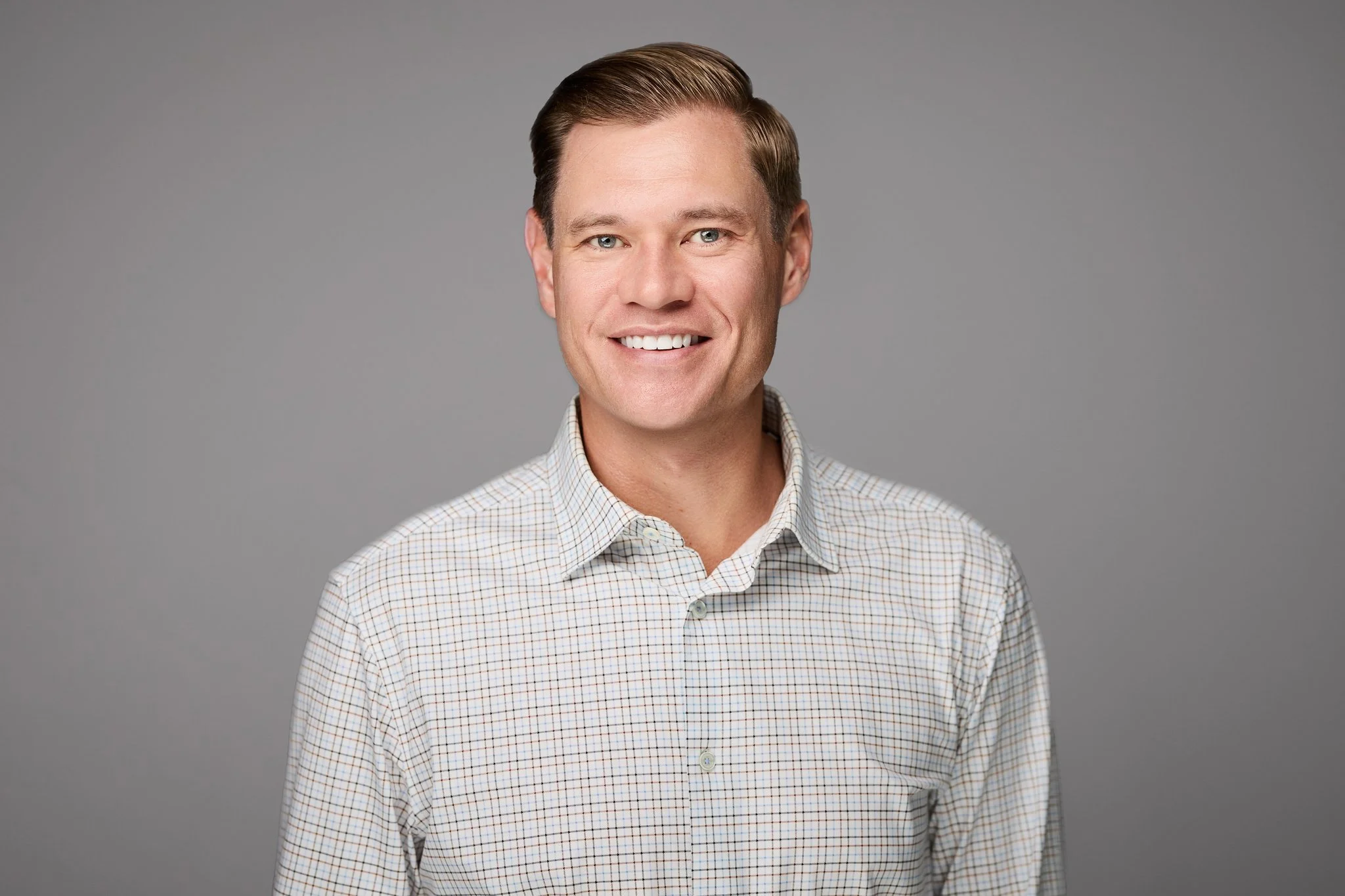 Professional headshot of a man with brown hair, blue eyes, smiling, wearing a light-colored checkered shirt, against a plain gray background.
