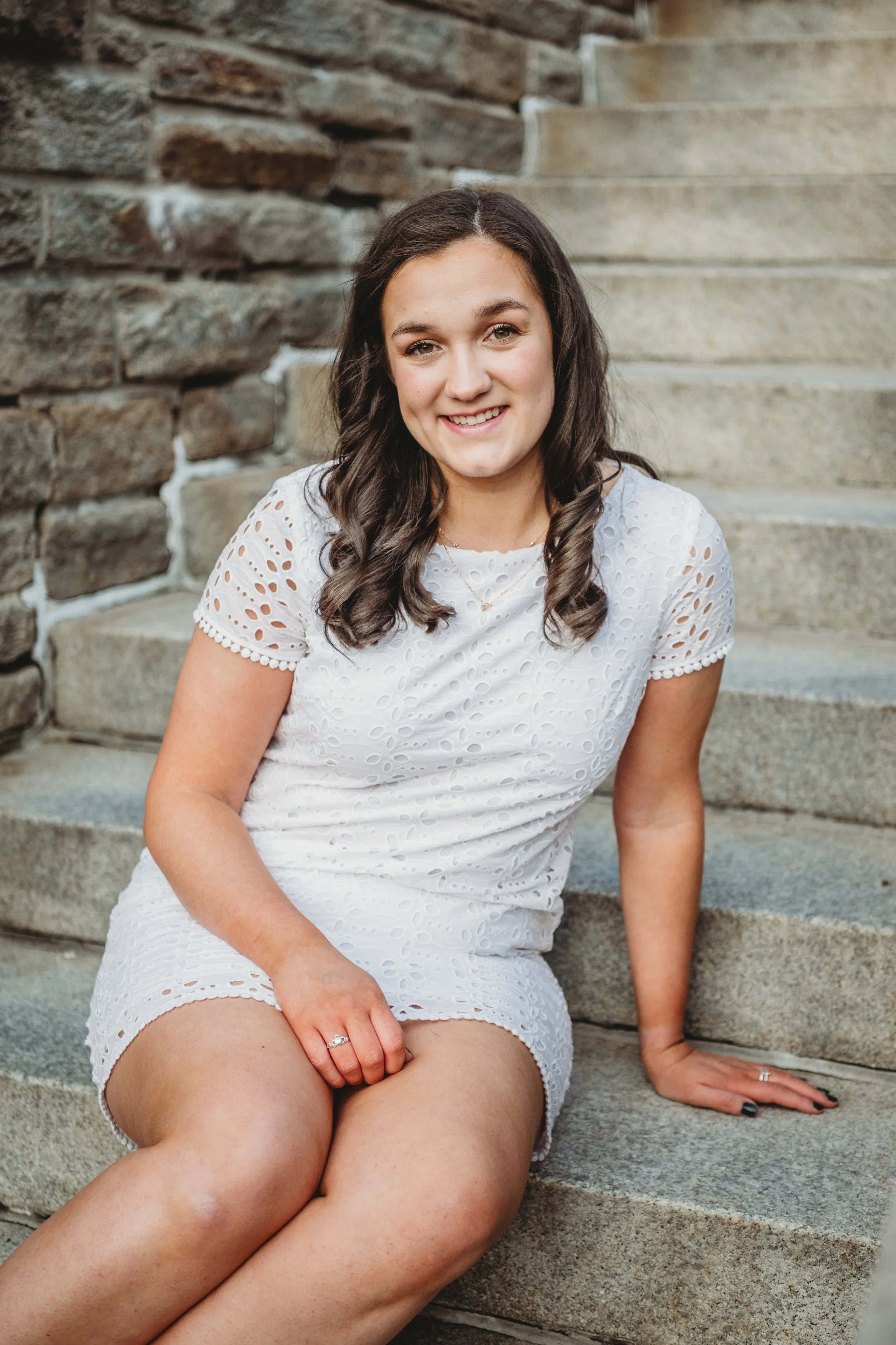 A young woman with wavy dark hair sitting on stone steps, smiling, wearing a white eyelet dress with short sleeves.
