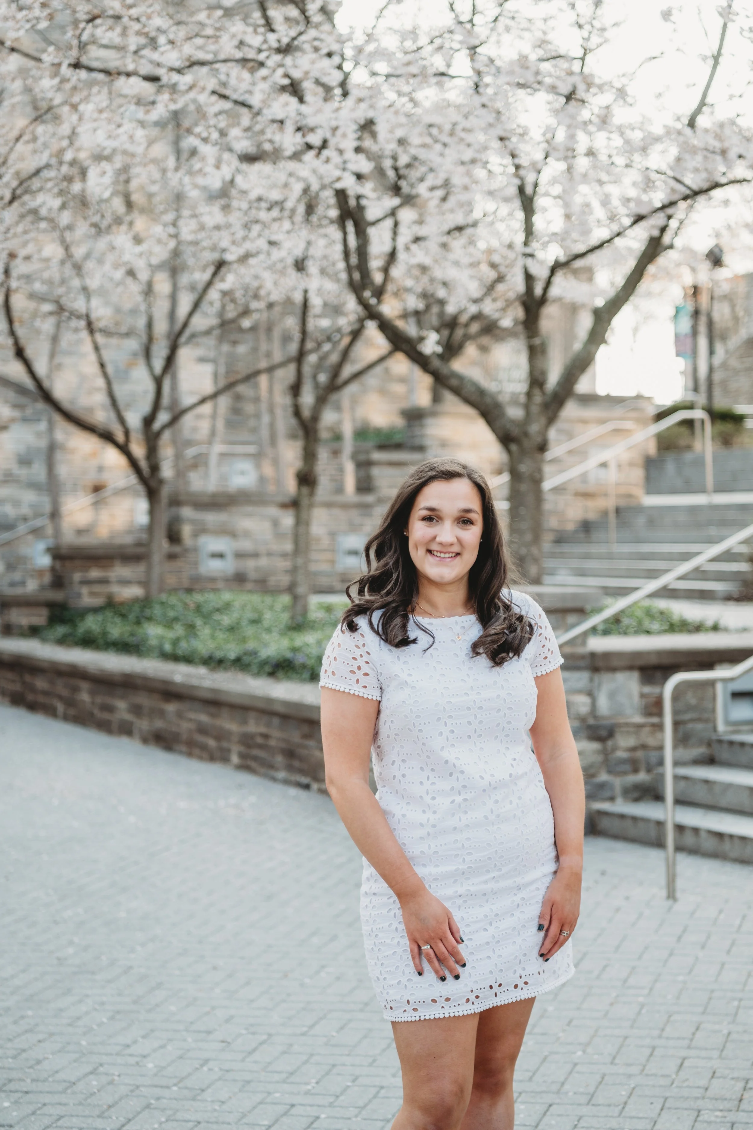 A smiling woman in a white eyelet dress standing outdoors in front of flowering trees and stone steps.