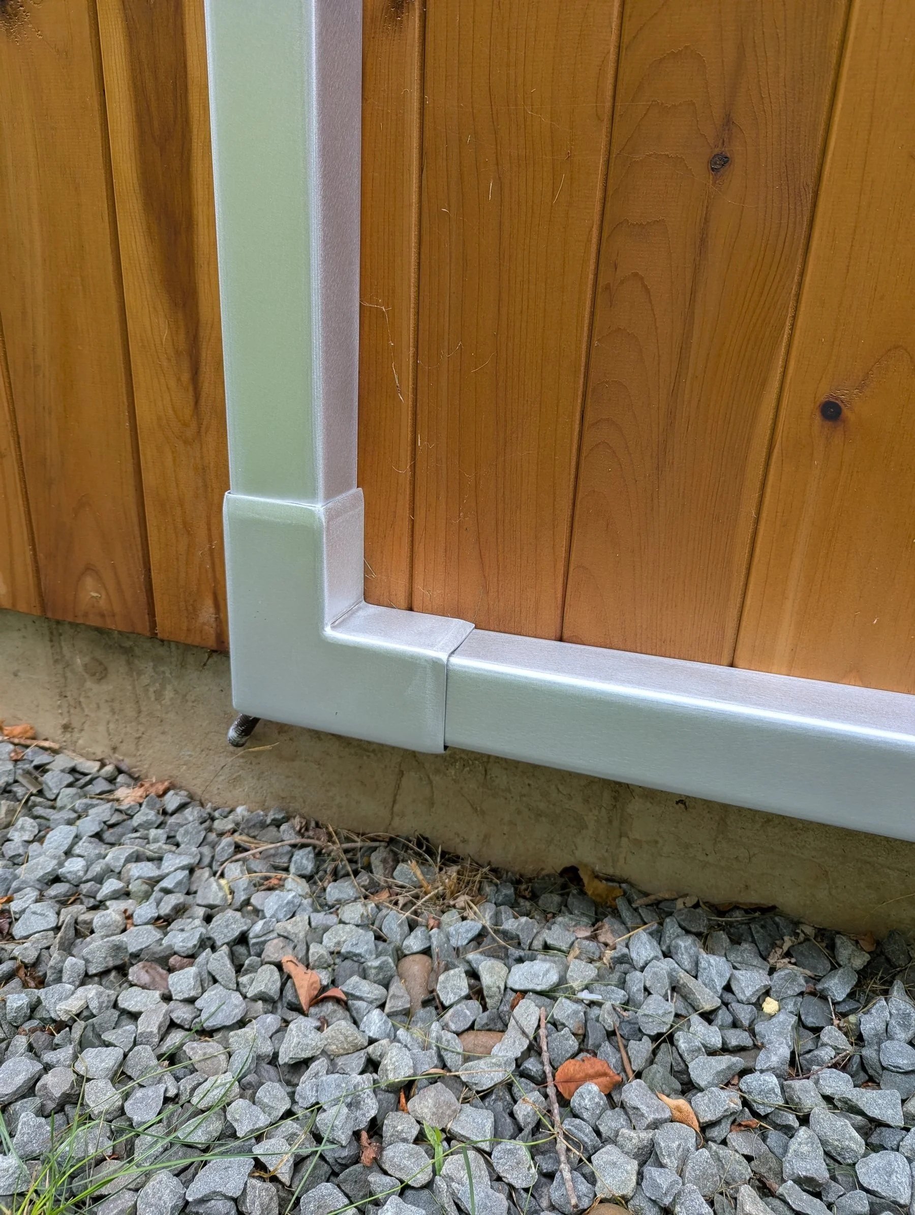 Close-up of the corner of a wooden building, showing a concrete foundation and gravel ground with scattered leaves and grass.