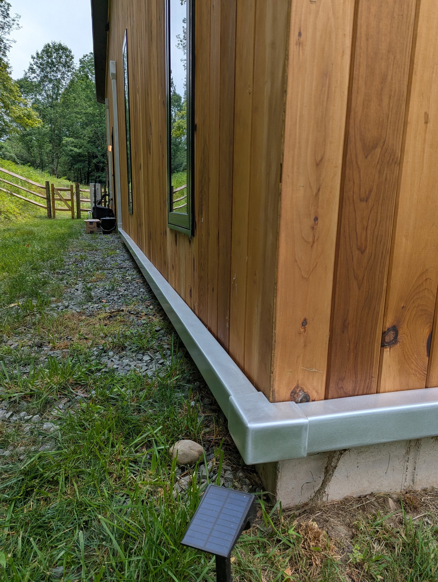 Side view of a wooden house exterior with orbital sanded aluminum mini split line cover, two windows, and a small solar panel on the ground nearby, surrounded by grass and trees.