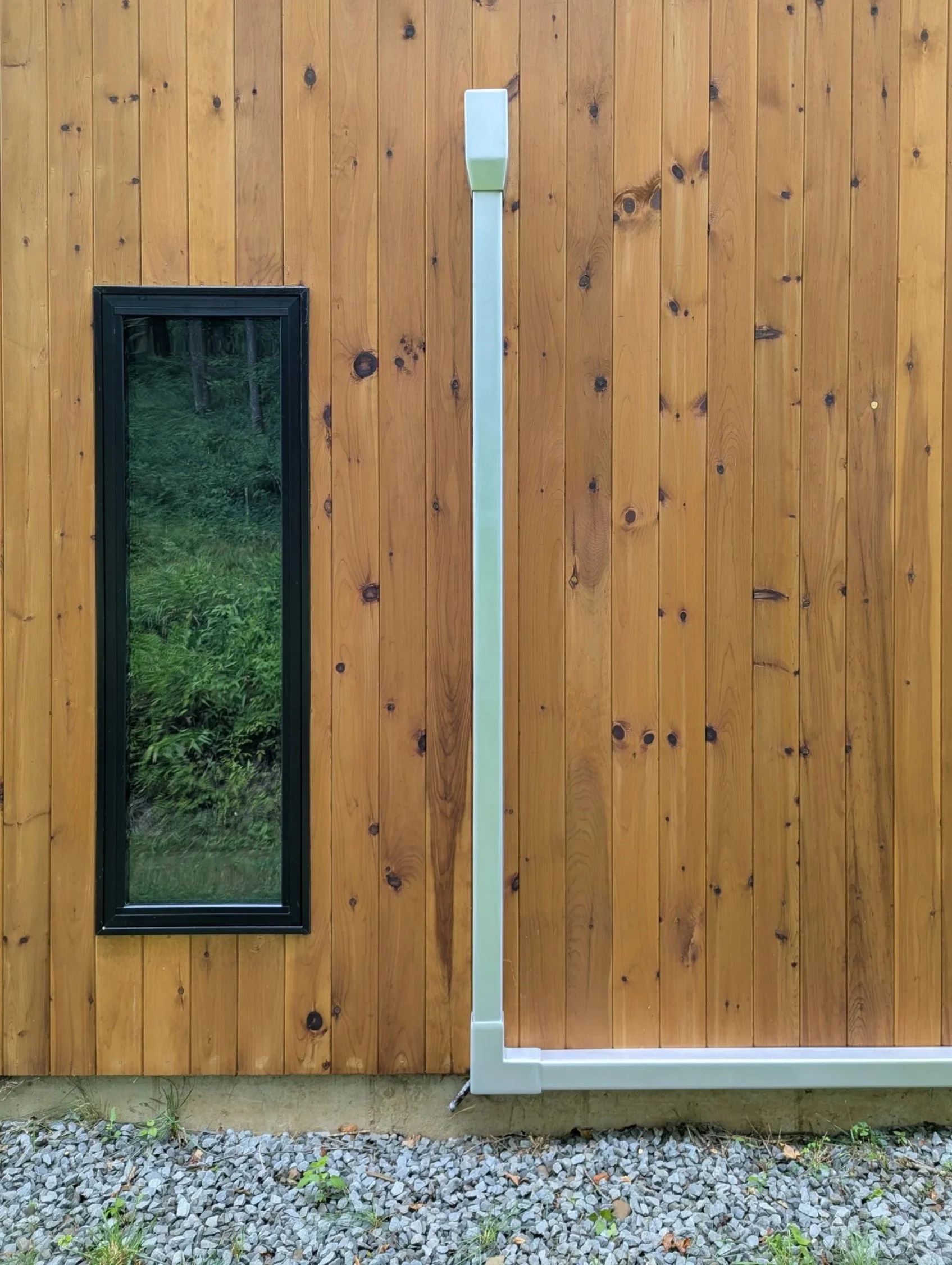 A wooden exterior wall of a building with a small rectangular window on the left and a tall, orbital sanded aluminum mini split line cover. The ground below has small rocks and some green plants.