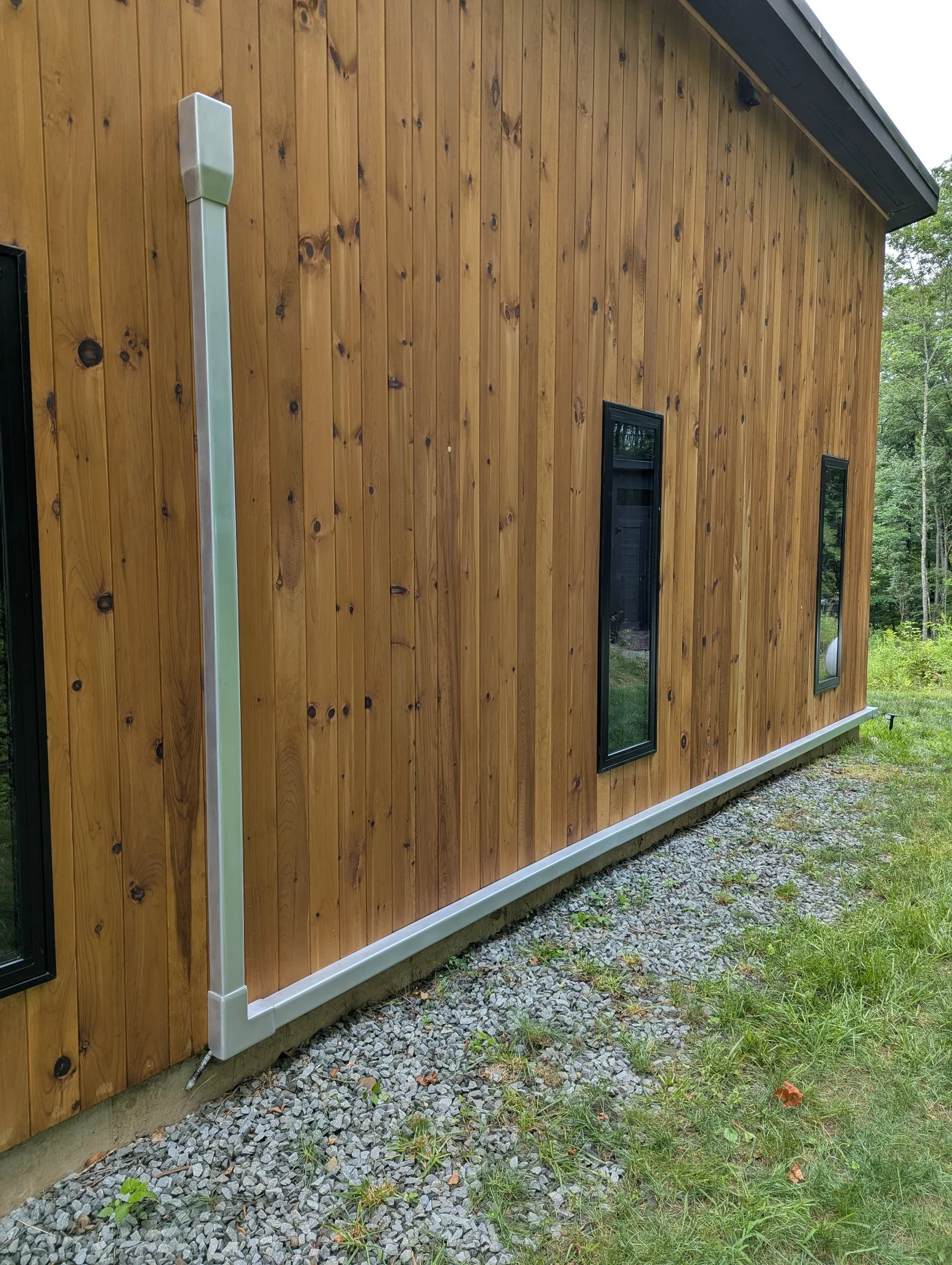 The exterior of a wooden building with vertical wood paneling, two small rectangular windows, orbital sanded aluminum mini split line cover and a downspout. The ground is covered with gravel and some grass.