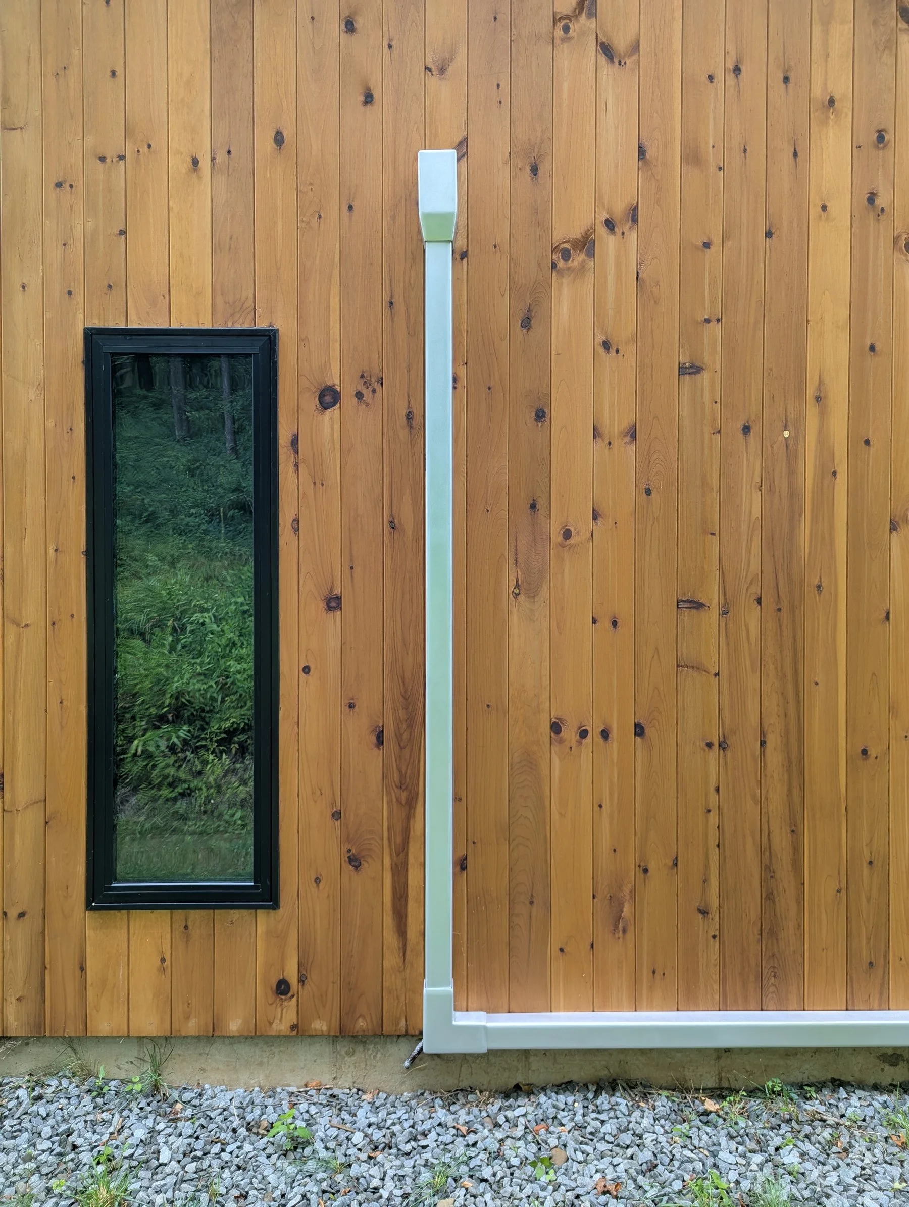 The image shows a wooden wall with a small rectangular window on the left side and orbital sanded aluminum mini split line cover along the middle and bottom, running vertically and horizontally, on the right side. 