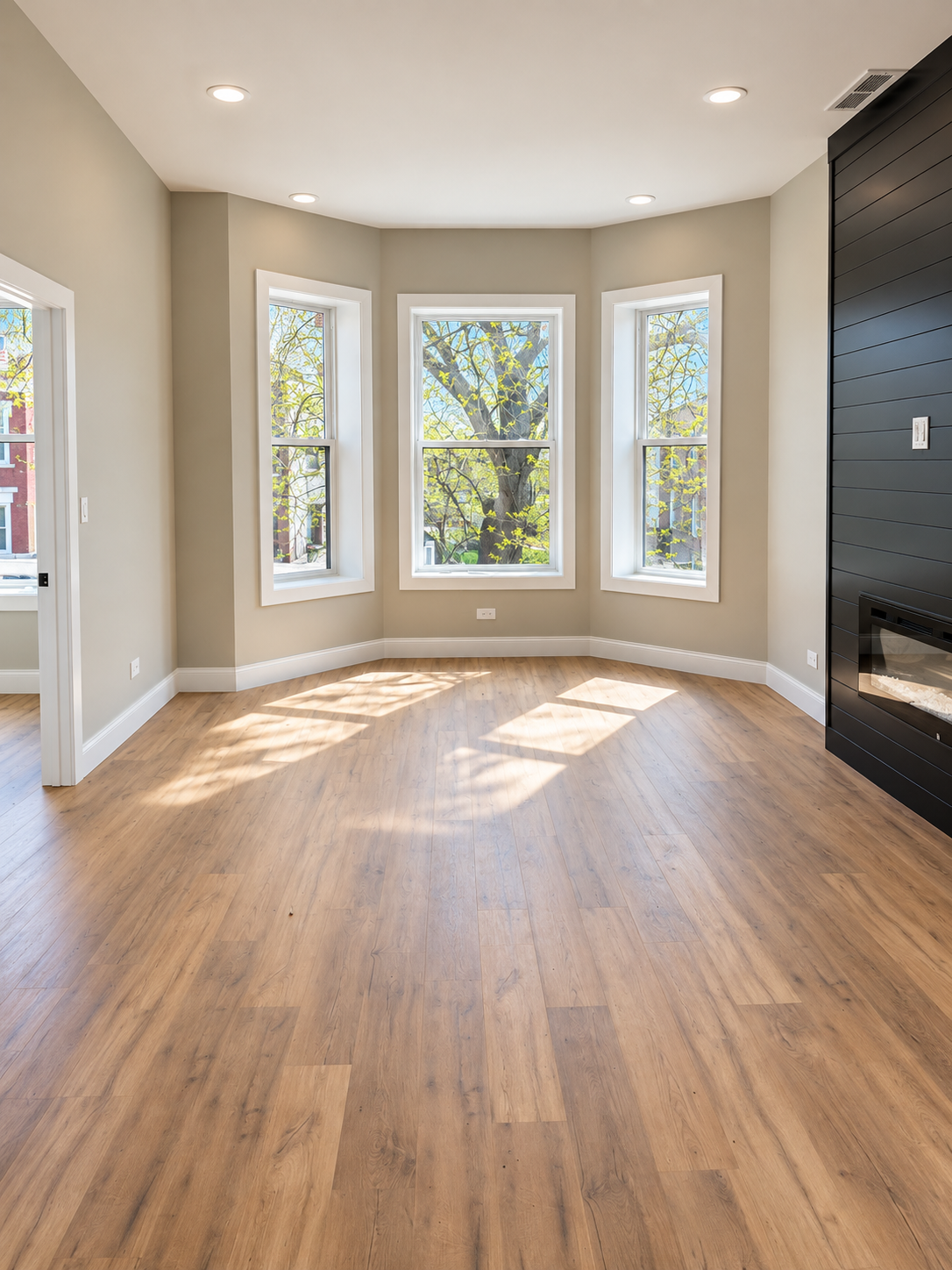 Empty living room with three large windows, hardwood floors, and a black fireplace on the right wall, illuminated by ceiling lights.