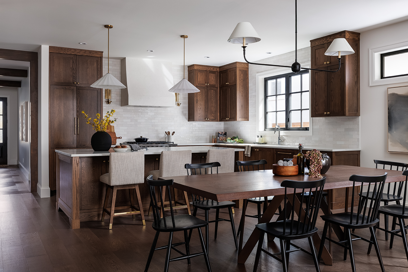 Modern kitchen with dark wood cabinets, white walls, and a wooden dining table surrounded by black chairs and beige upholstered barstools, with decorative items on the table.