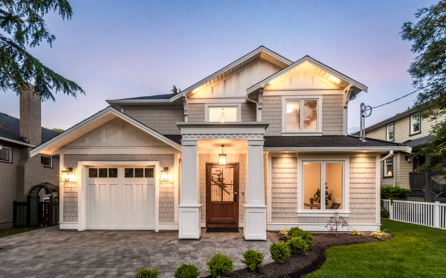 A two-story suburban house with beige siding, white trim, a front porch with a wooden door, a attached garage, and a well-maintained garden with small shrubs and grass, illuminated at dusk.