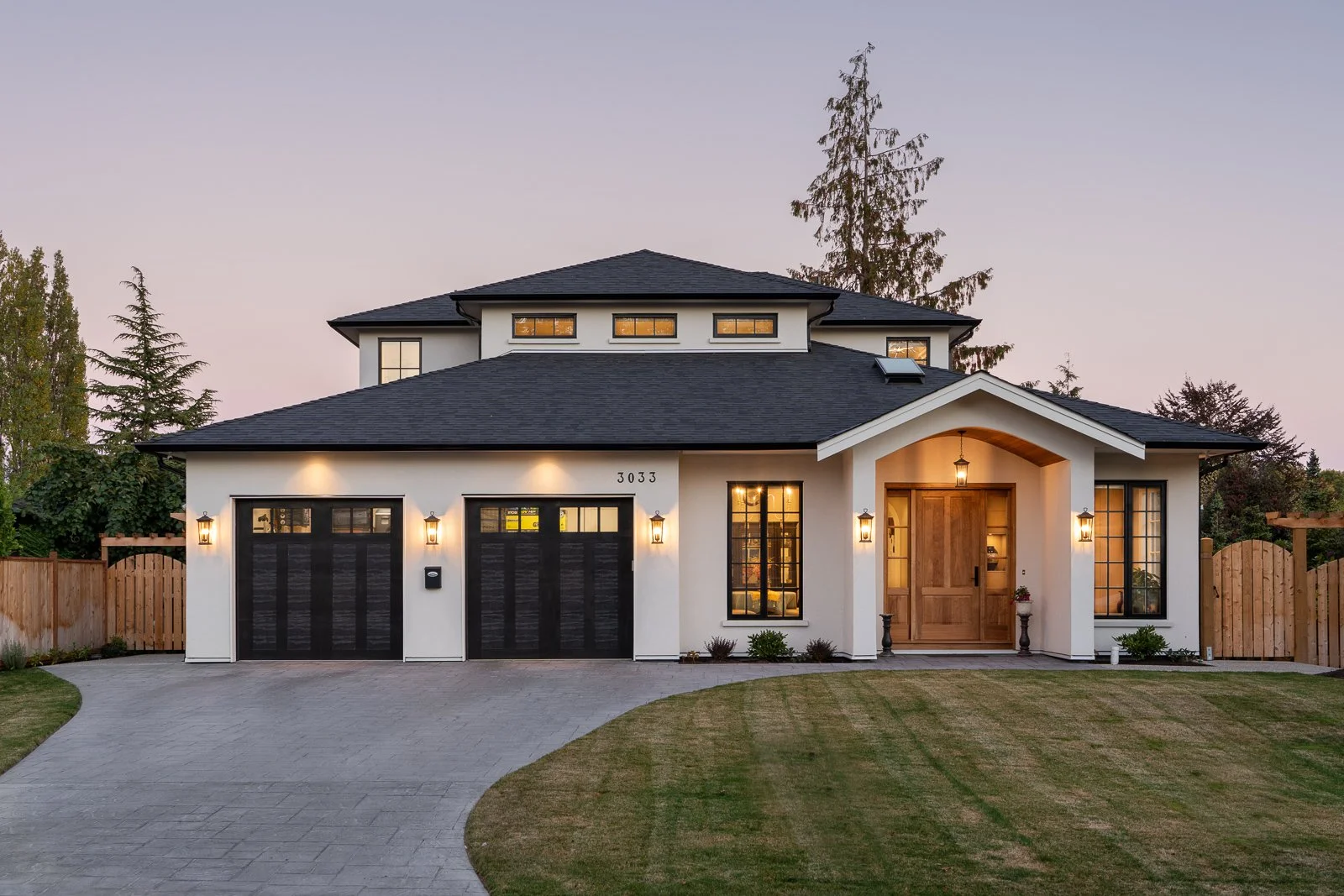 Front view of a modern house with a curved driveway, fenced yard, two-car garage, wooden front door, and exterior lights during dusk