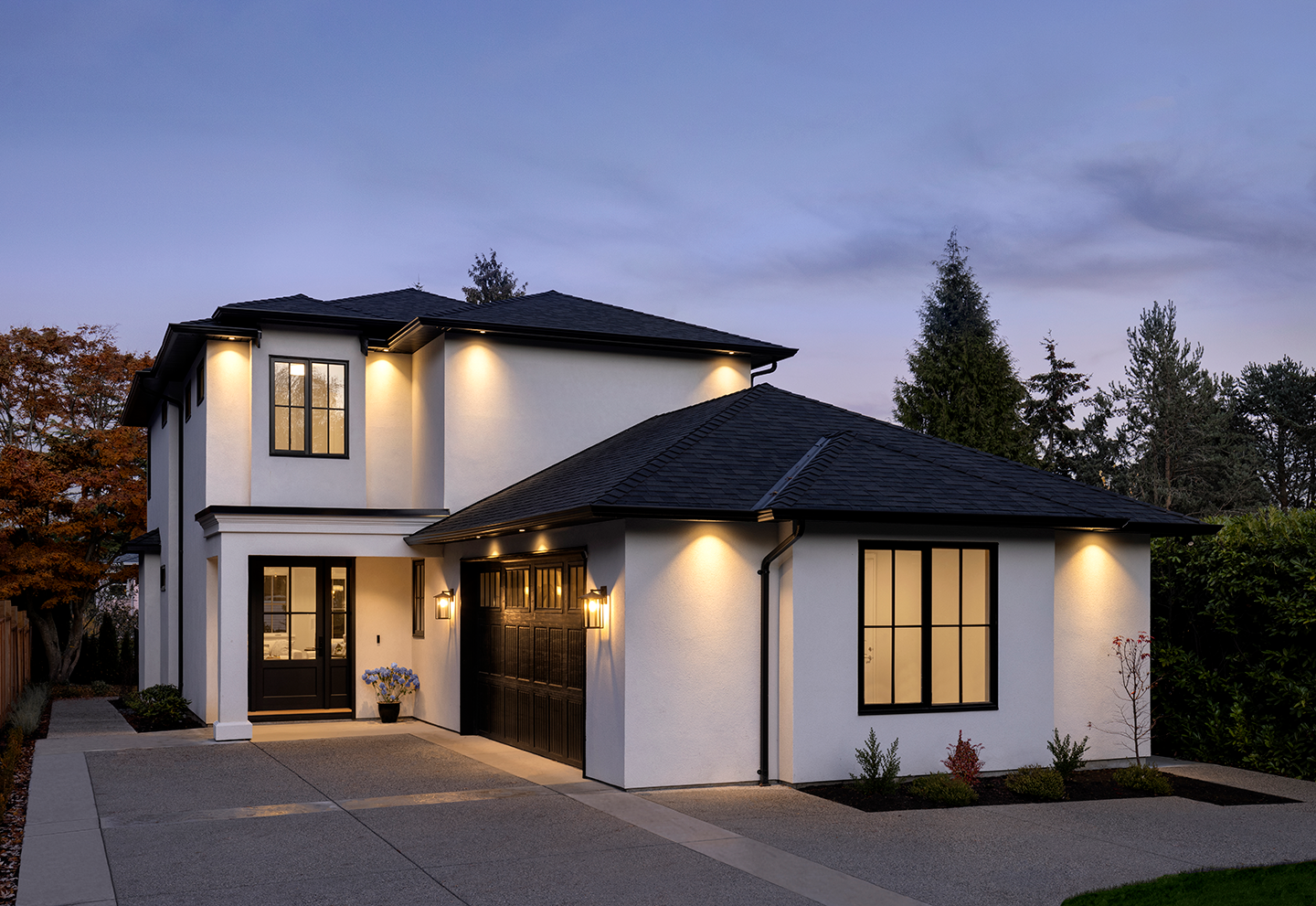 Modern two-story house with white exterior walls, black roof, black window frames, and front door, illuminated with outdoor lighting at dusk.