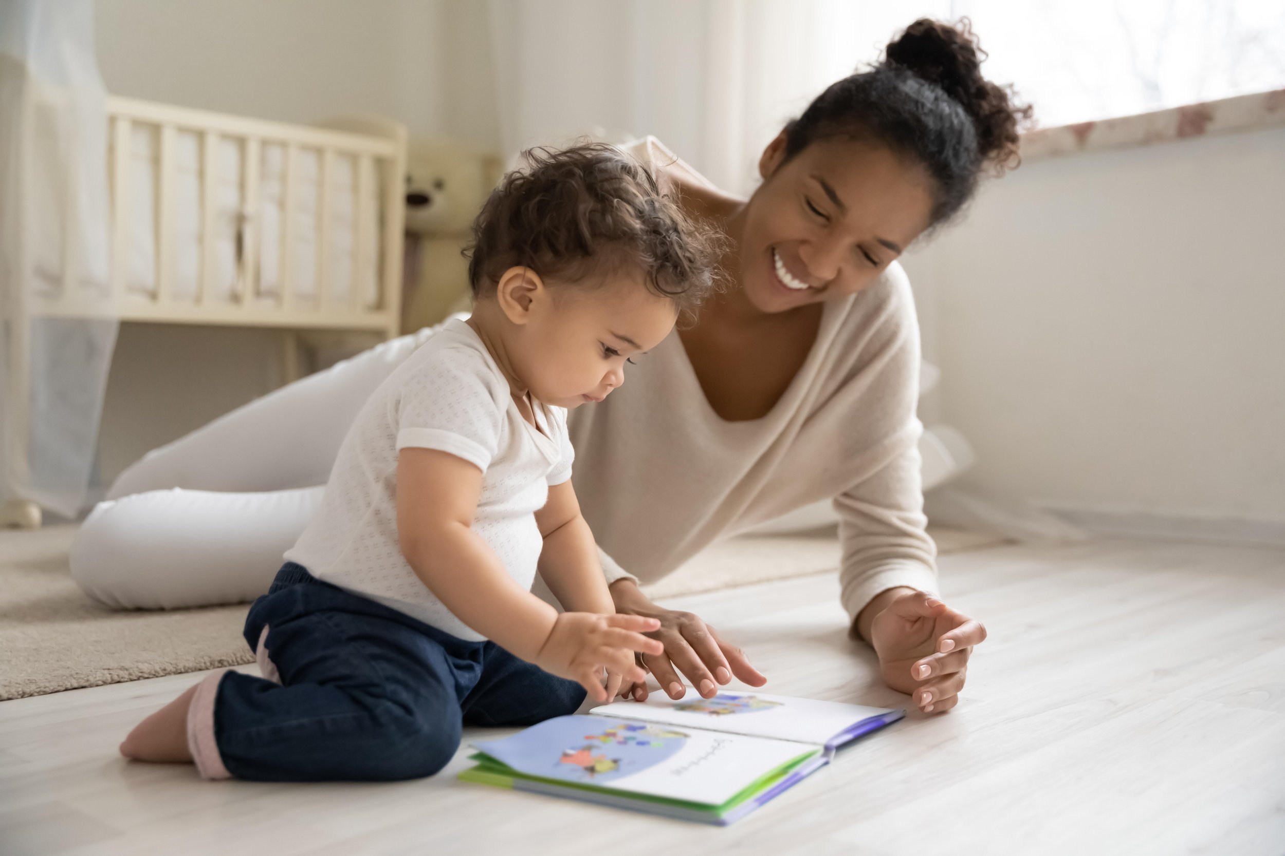 A woman and a young child sitting on the floor together, engaging in a puzzle or activity with colorful pieces. They are smiling and appear to be enjoying their time indoors near a window with natural light.