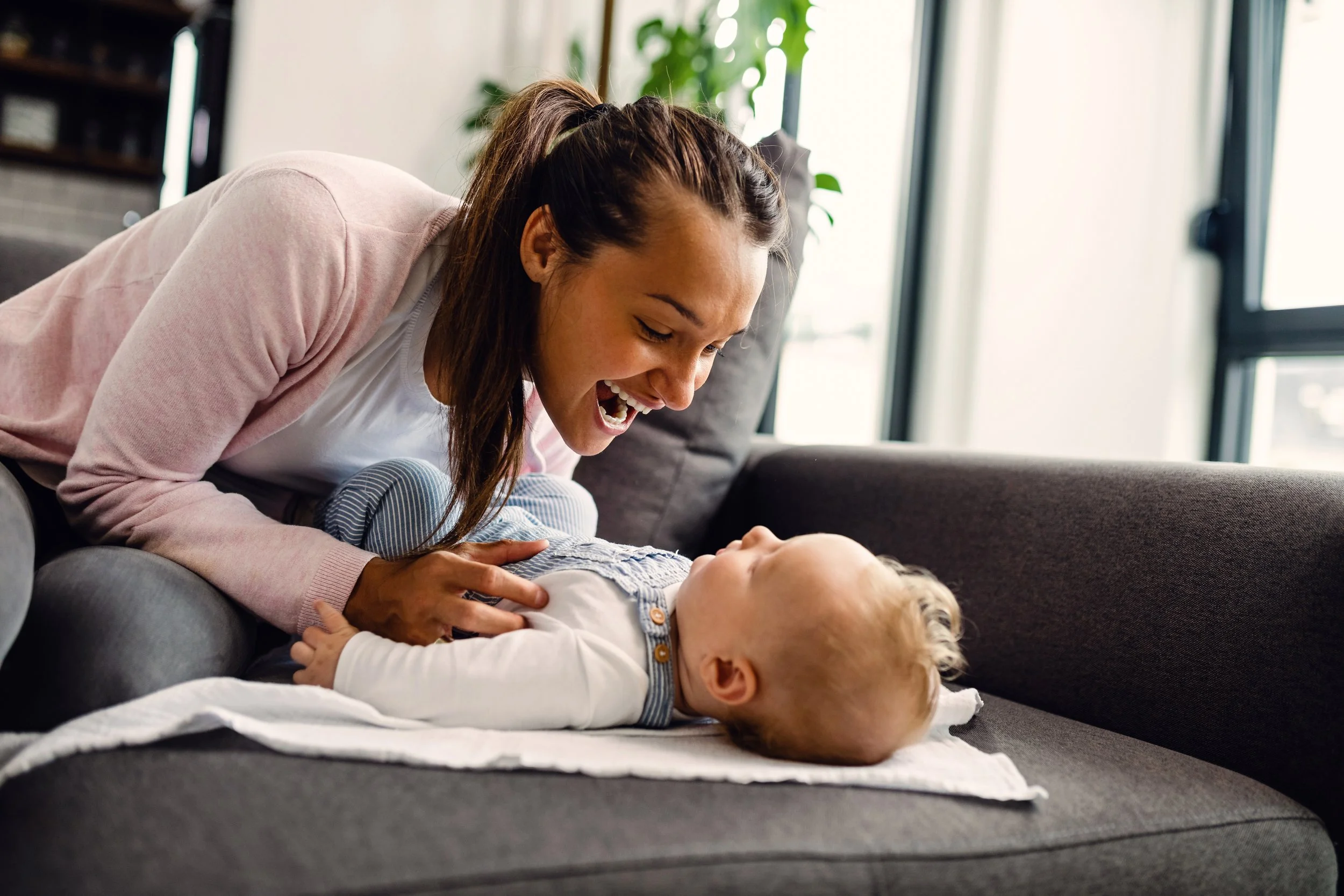 A woman and a baby on a gray couch, laughing and playing together in a bright living room with large windows.
