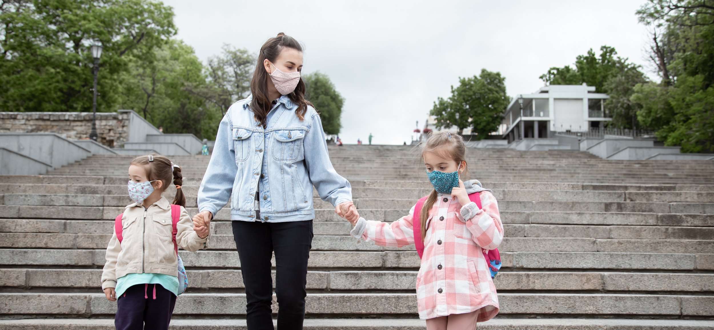 A woman holding hands with two young girls while walking down outdoor steps, all wearing face masks, with green trees and buildings in the background.